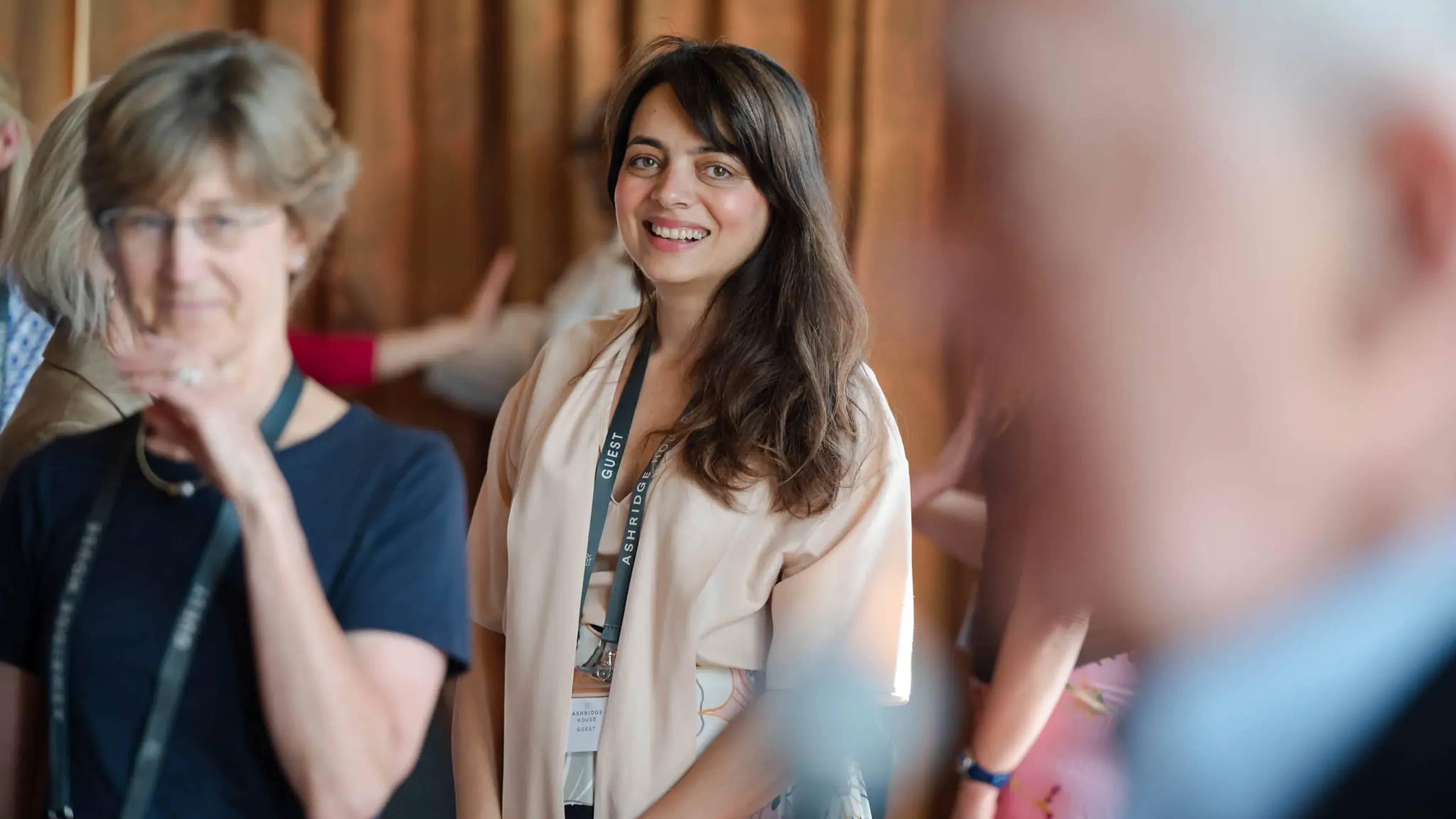 Smiling woman with a guest lanyard stands amidst a group, people blurred in the foreground and background, in a warmly lit room.