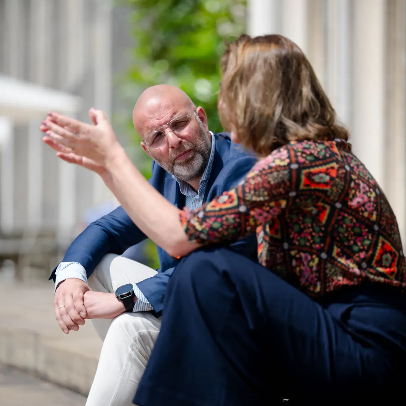 Two people sit outdoors, engaged in conversation. One gestures with their hand while the other listens attentively in a casual setting.