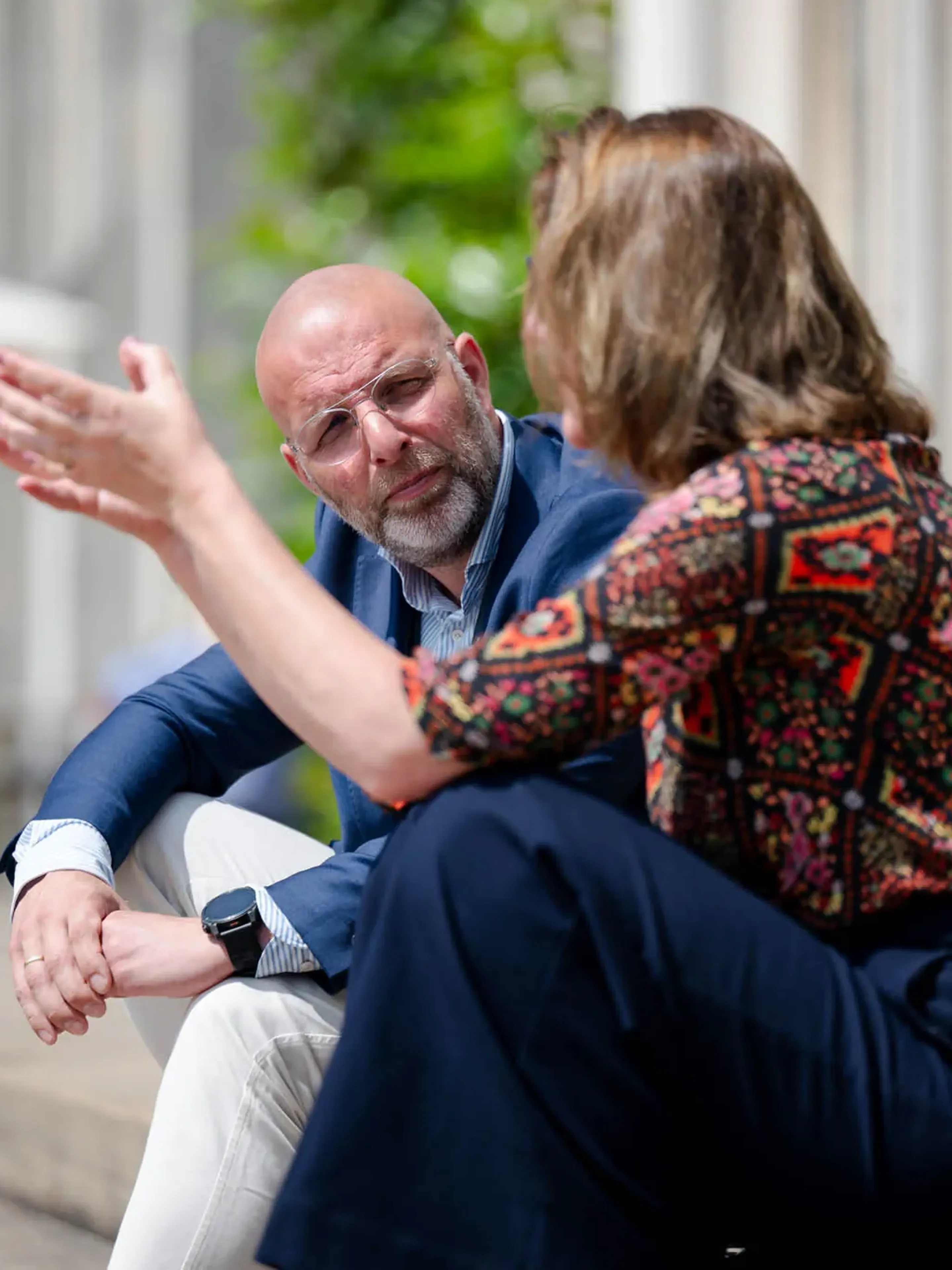 Two people sit outdoors, engaged in conversation. One gestures with their hand while the other listens attentively in a casual setting.