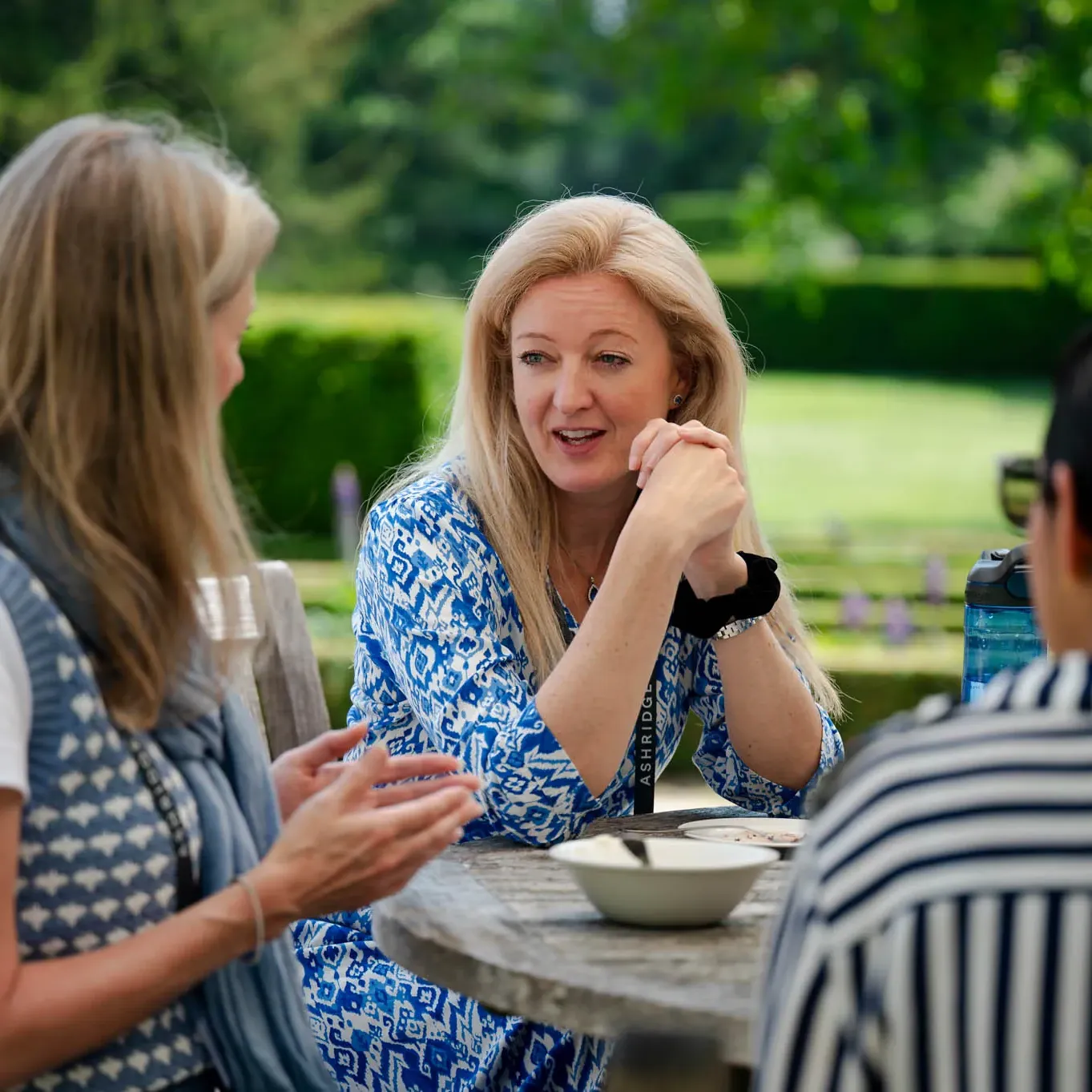 Three women sit at a round wooden table outdoors, engaged in conversation, surrounded by greenery.