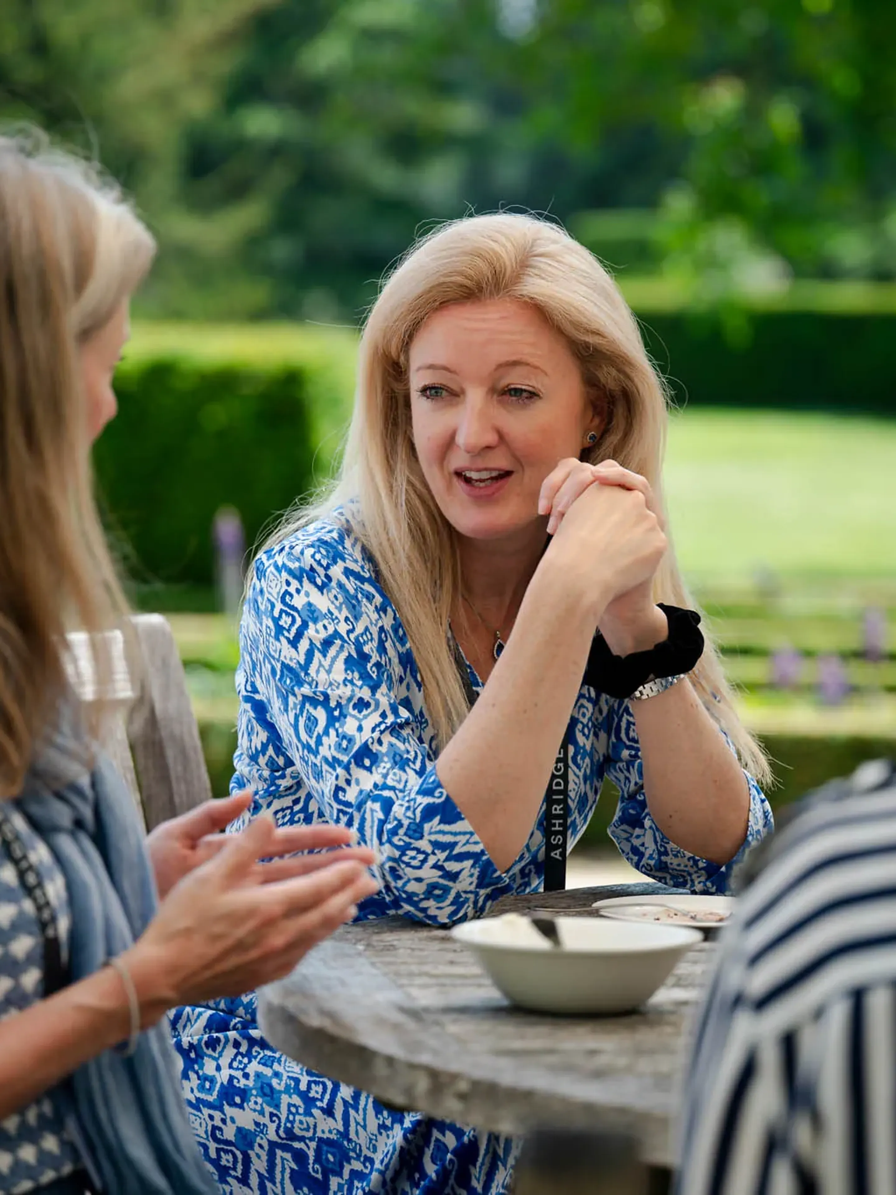 Three women sit at a round wooden table outdoors, engaged in conversation, surrounded by greenery.