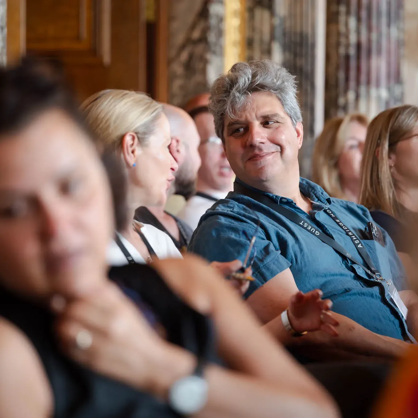 People seated in a conference room, engaged in conversation. One person in a blue shirt smiles, others listen attentively nearby.