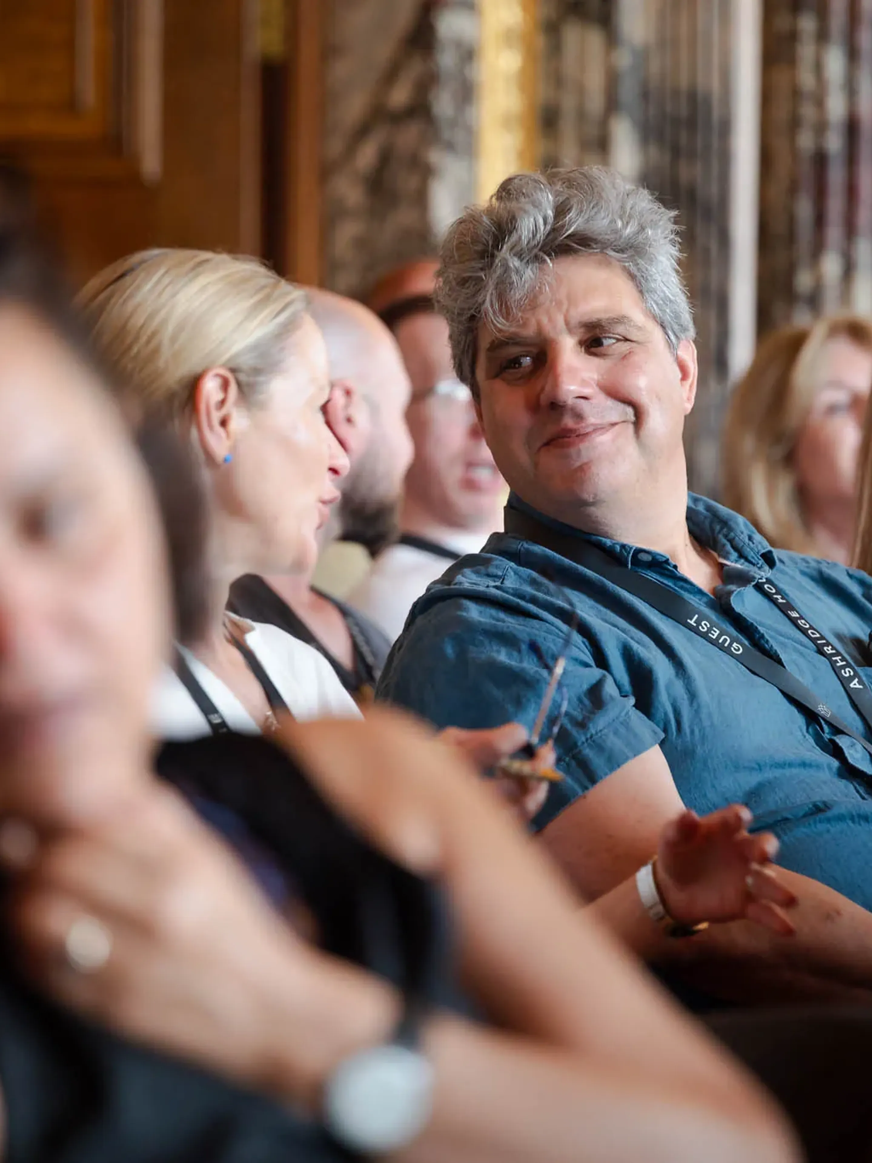 People seated in a conference room, engaged in conversation. One person in a blue shirt smiles, others listen attentively nearby.