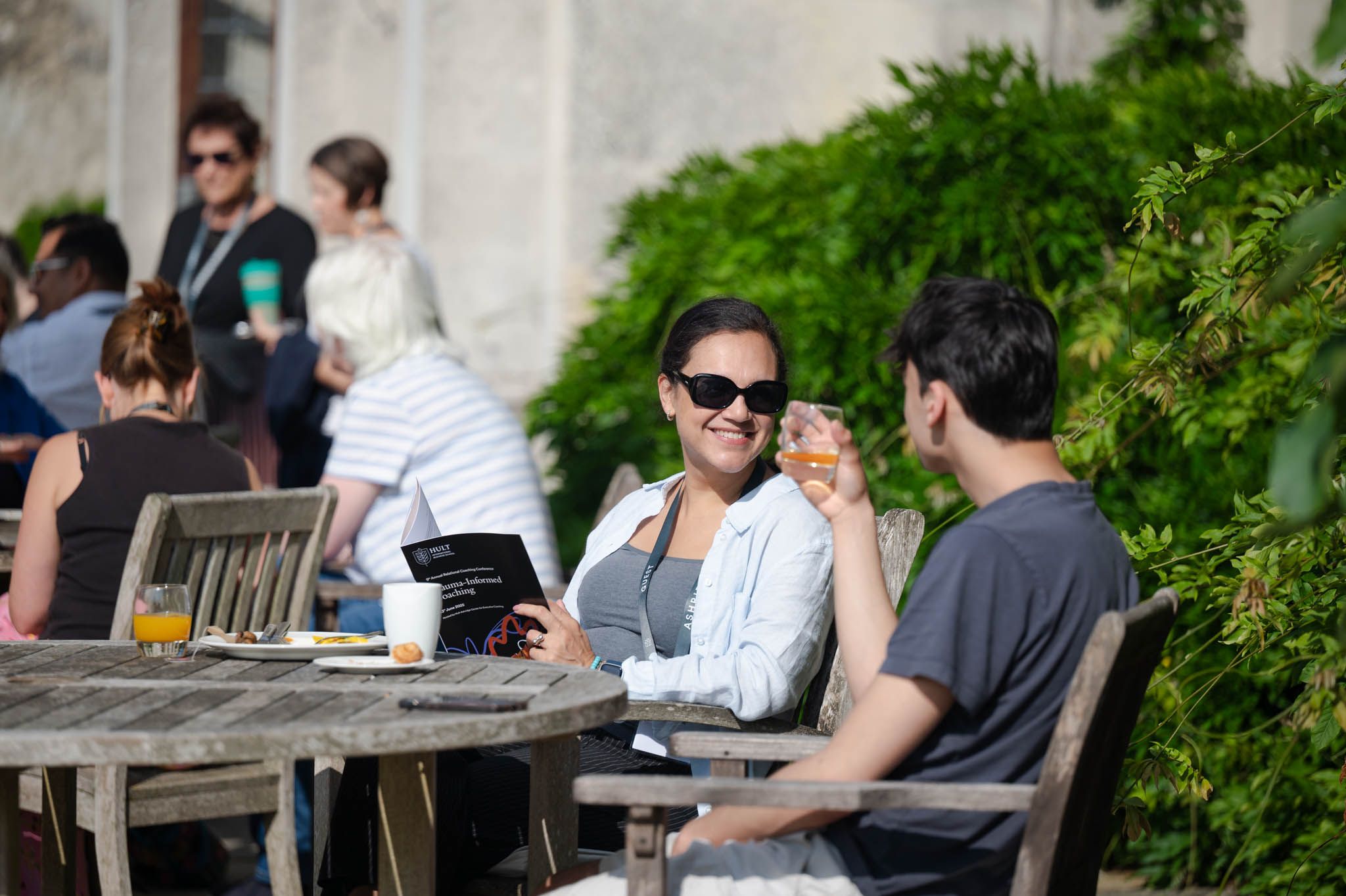 People seated outdoors at wooden tables, smiling and talking. A woman holds a booklet, and a man holds a drink. Greenery in the background.