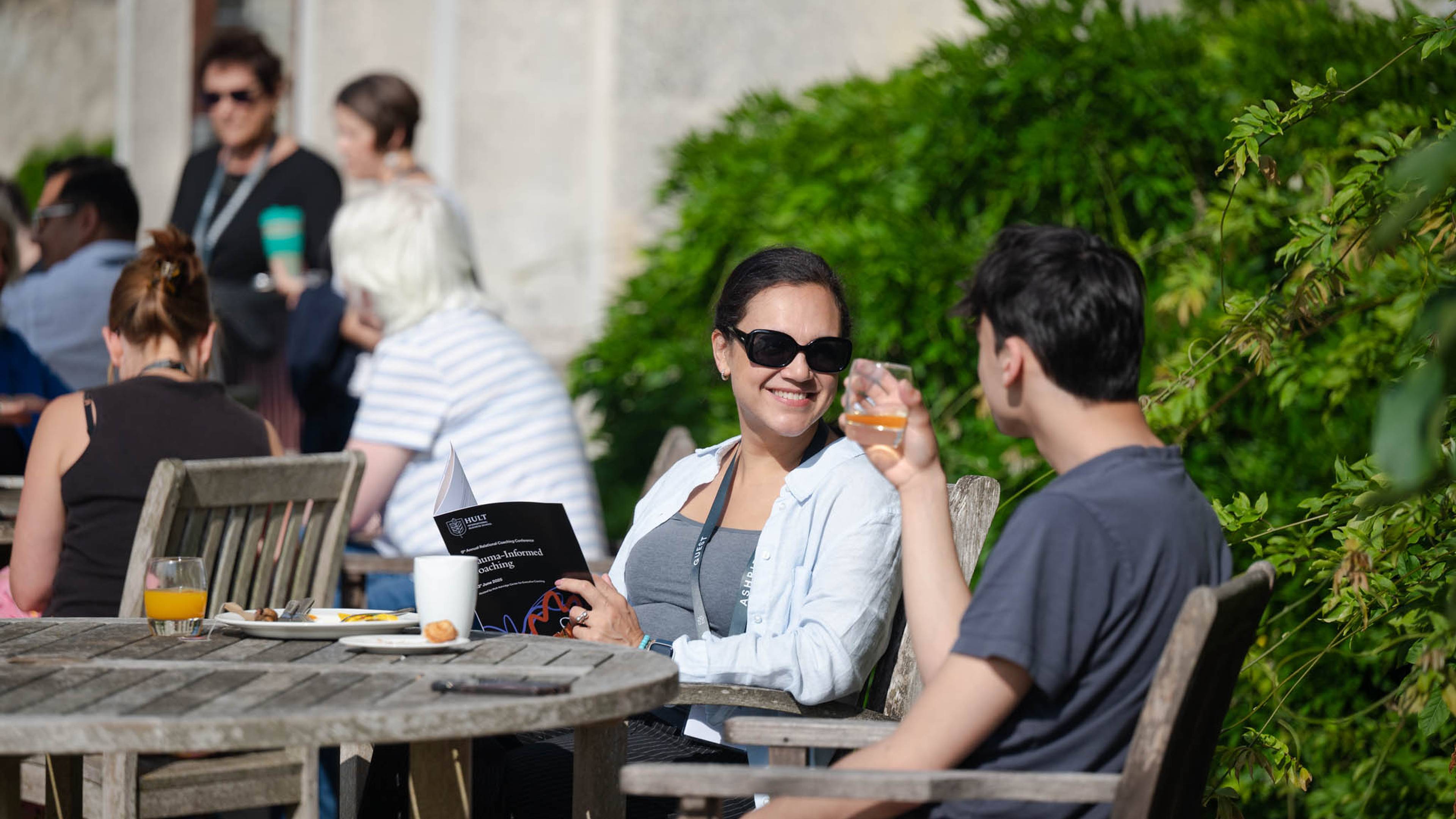 People seated outdoors at wooden tables, smiling and talking. A woman holds a booklet, and a man holds a drink. Greenery in the background.