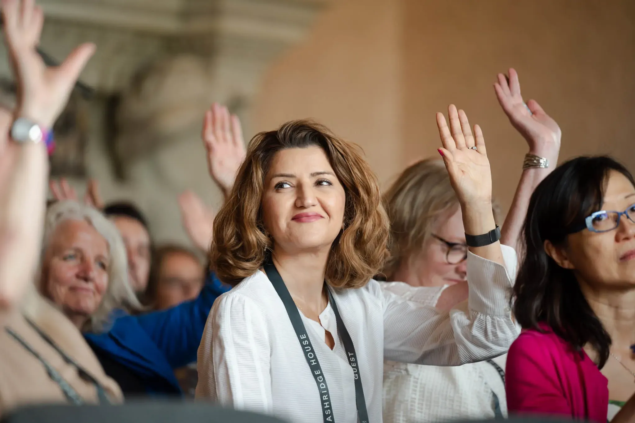 A group of people, predominantly women, raise their hands in what appears to be a conference or workshop setting.
