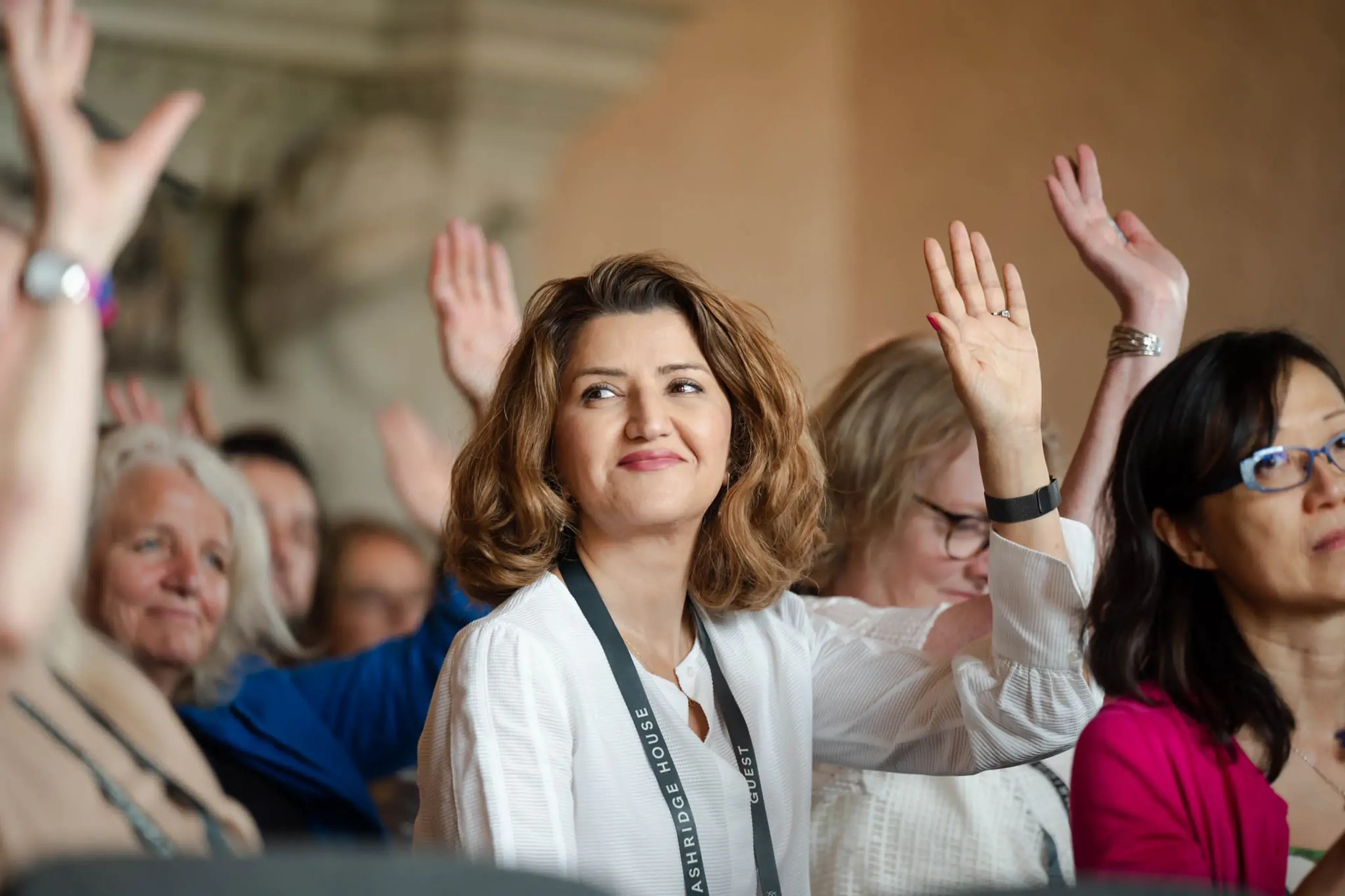 A group of people, predominantly women, raise their hands in what appears to be a conference or workshop setting.