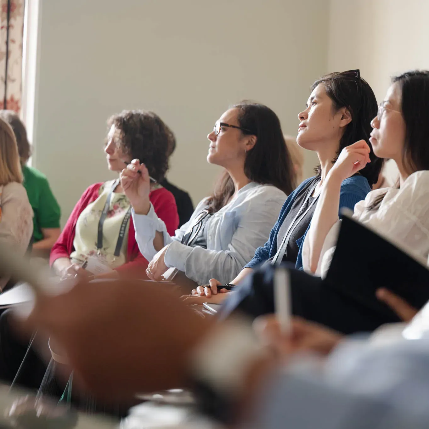 A diverse group of people attentively listens during a seminar, some taking notes.