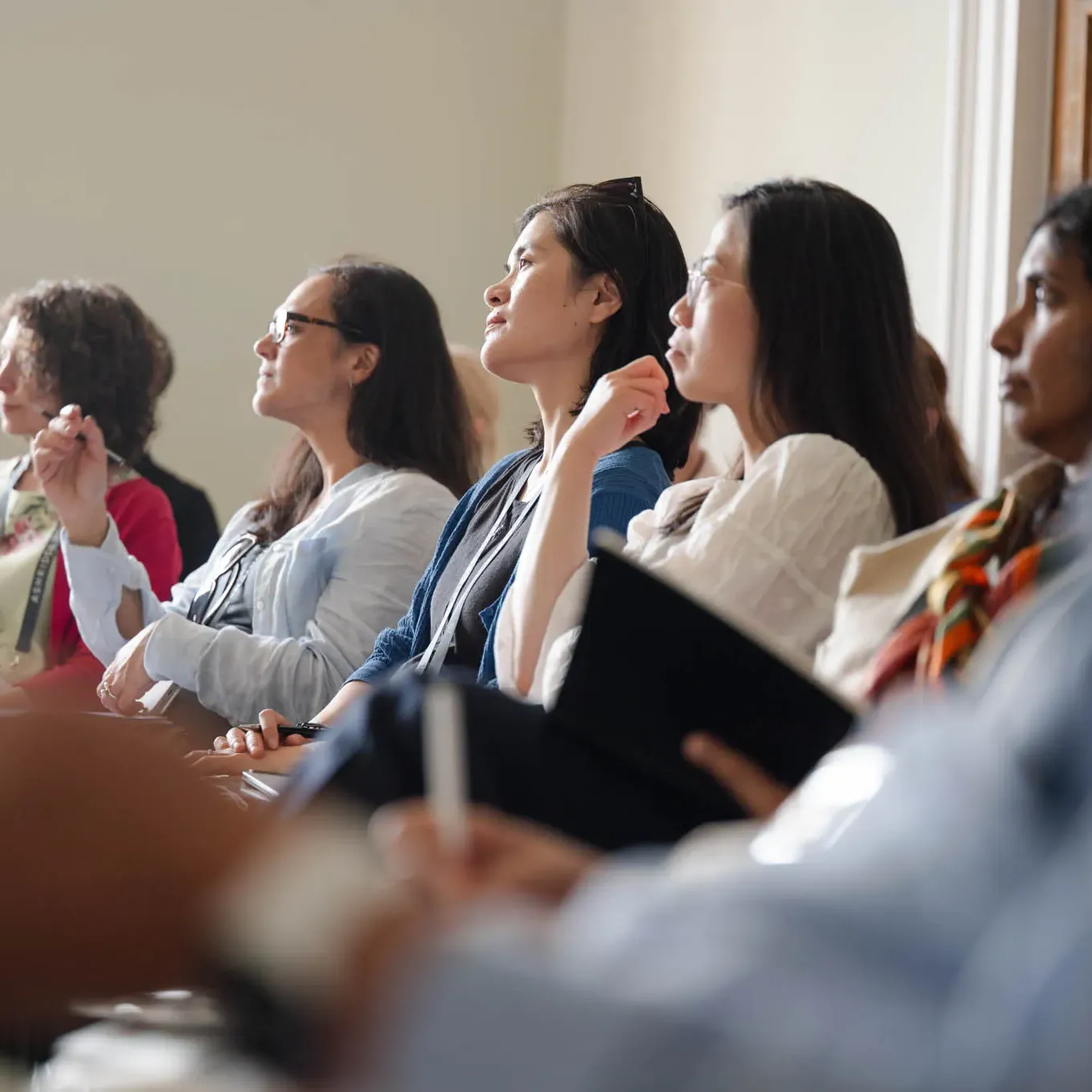 A diverse group of people attentively listens during a seminar, some taking notes.