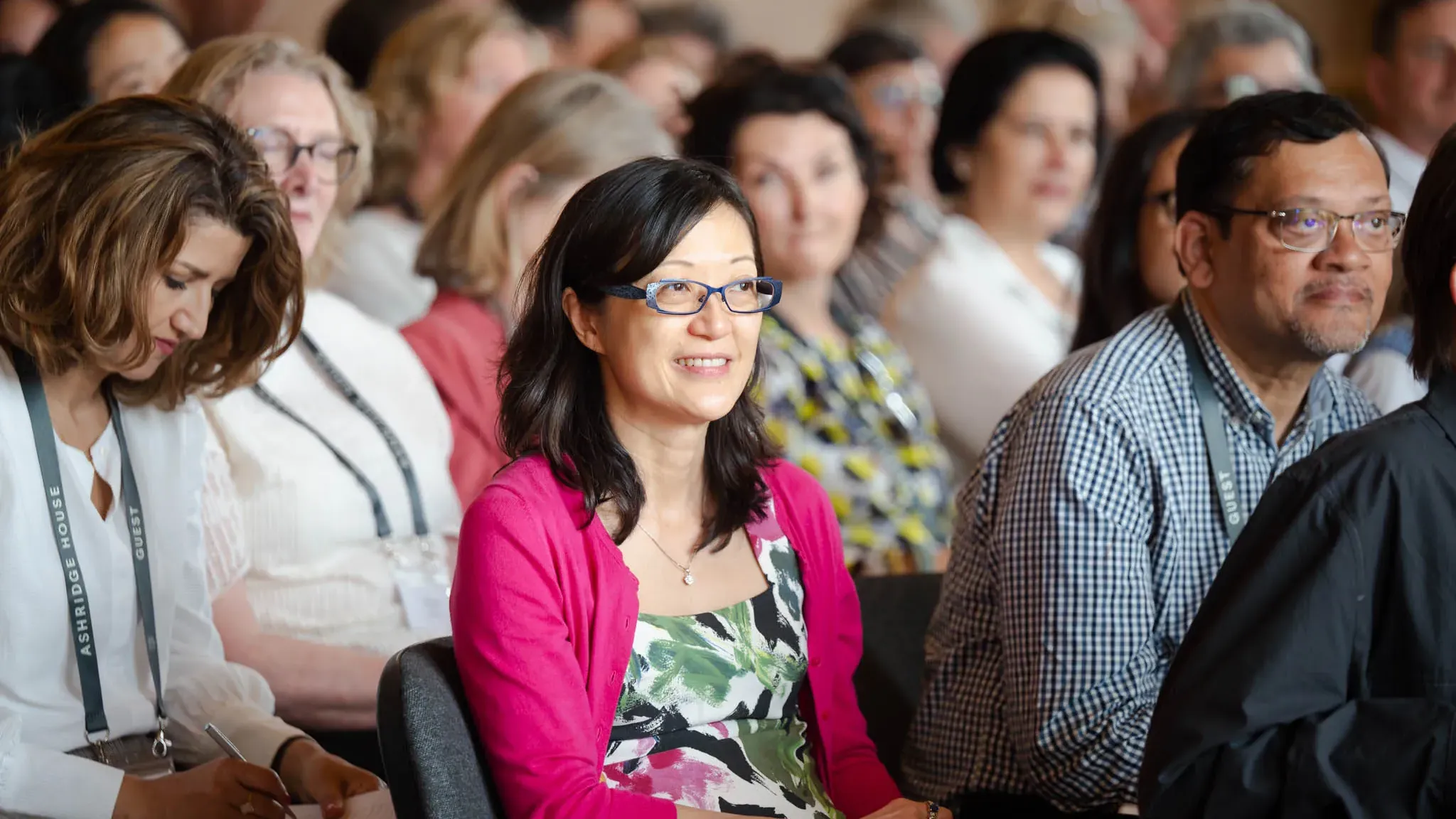 People seated in a conference room, attentively listening. A woman in a pink cardigan smiles, while another takes notes.
