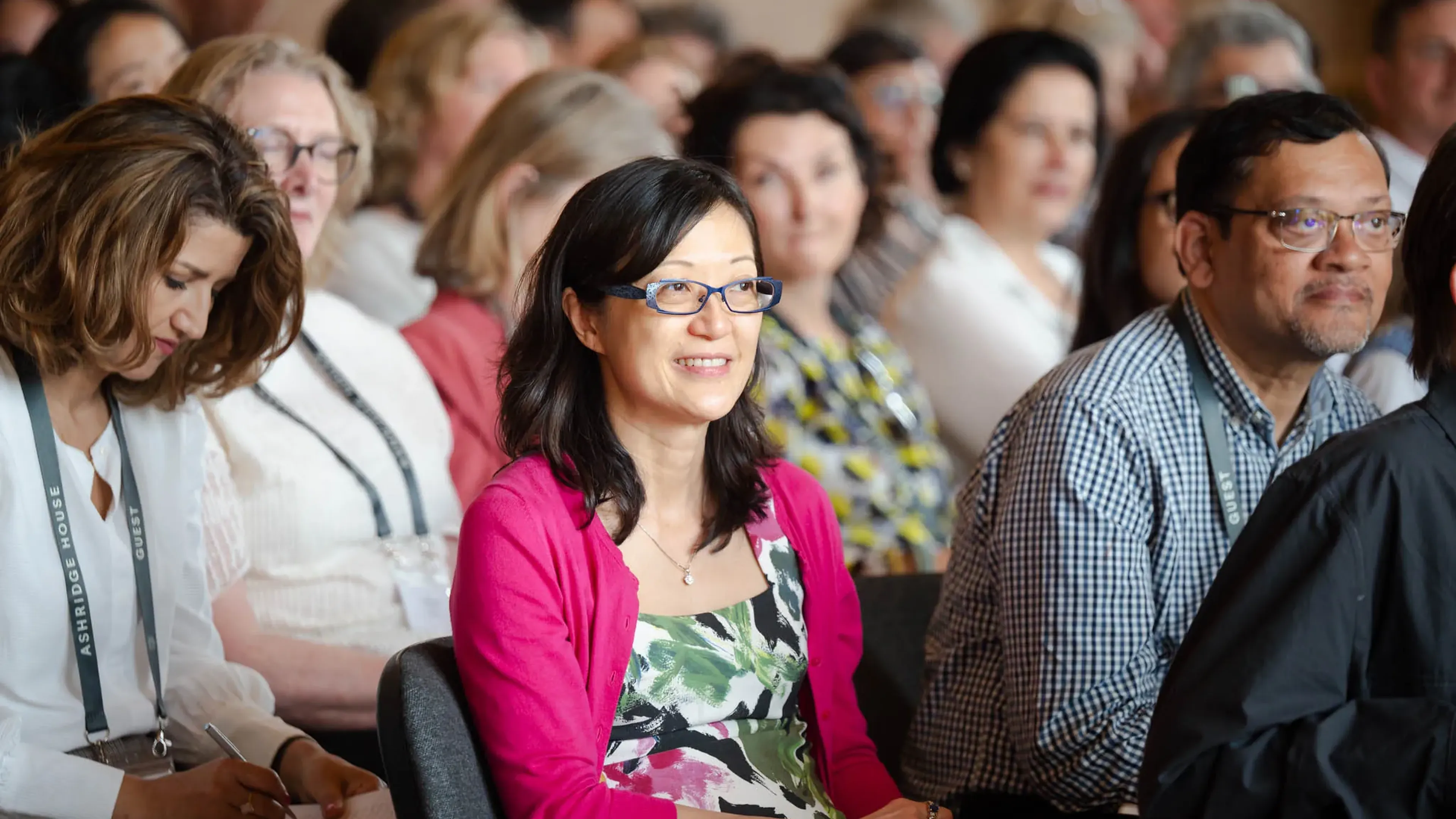 People seated in a conference room, attentively listening. A woman in a pink cardigan smiles, while another takes notes.
