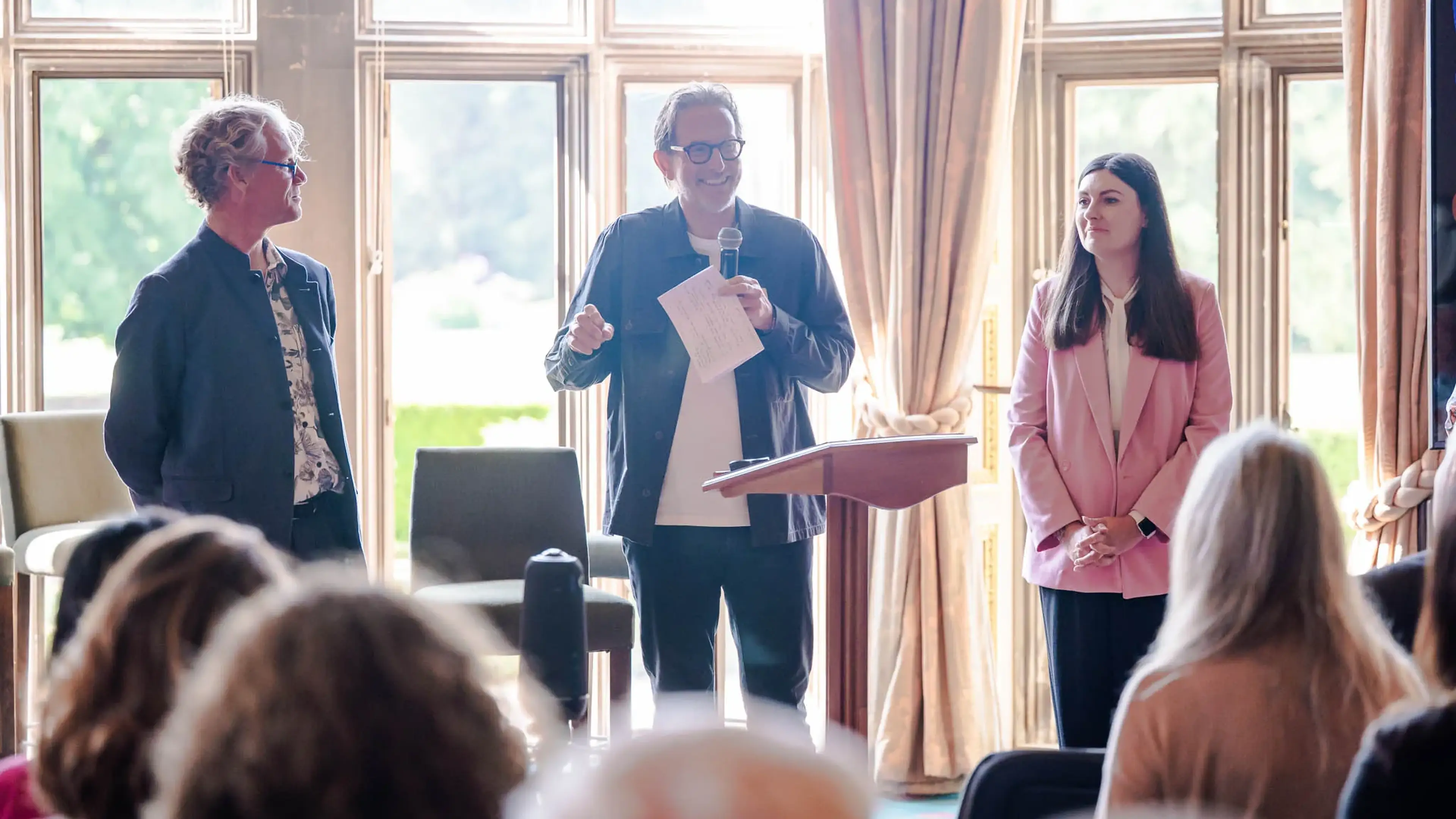 Three people stand in front of an audience in a bright room with large windows. One holds a microphone, smiling, as the others look on.