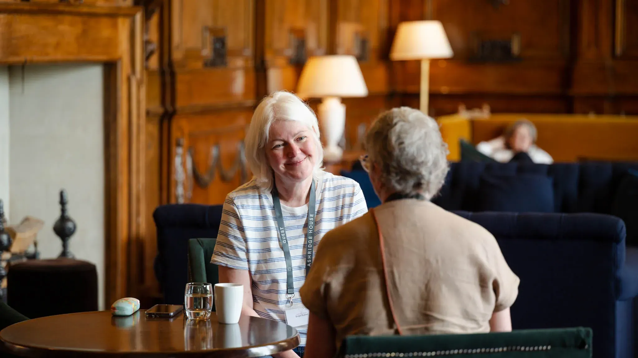 Two women sit at a wooden table in a cozy, wood-paneled room. One smiles warmly at the other, surrounded by comfortable seating and soft lighting.