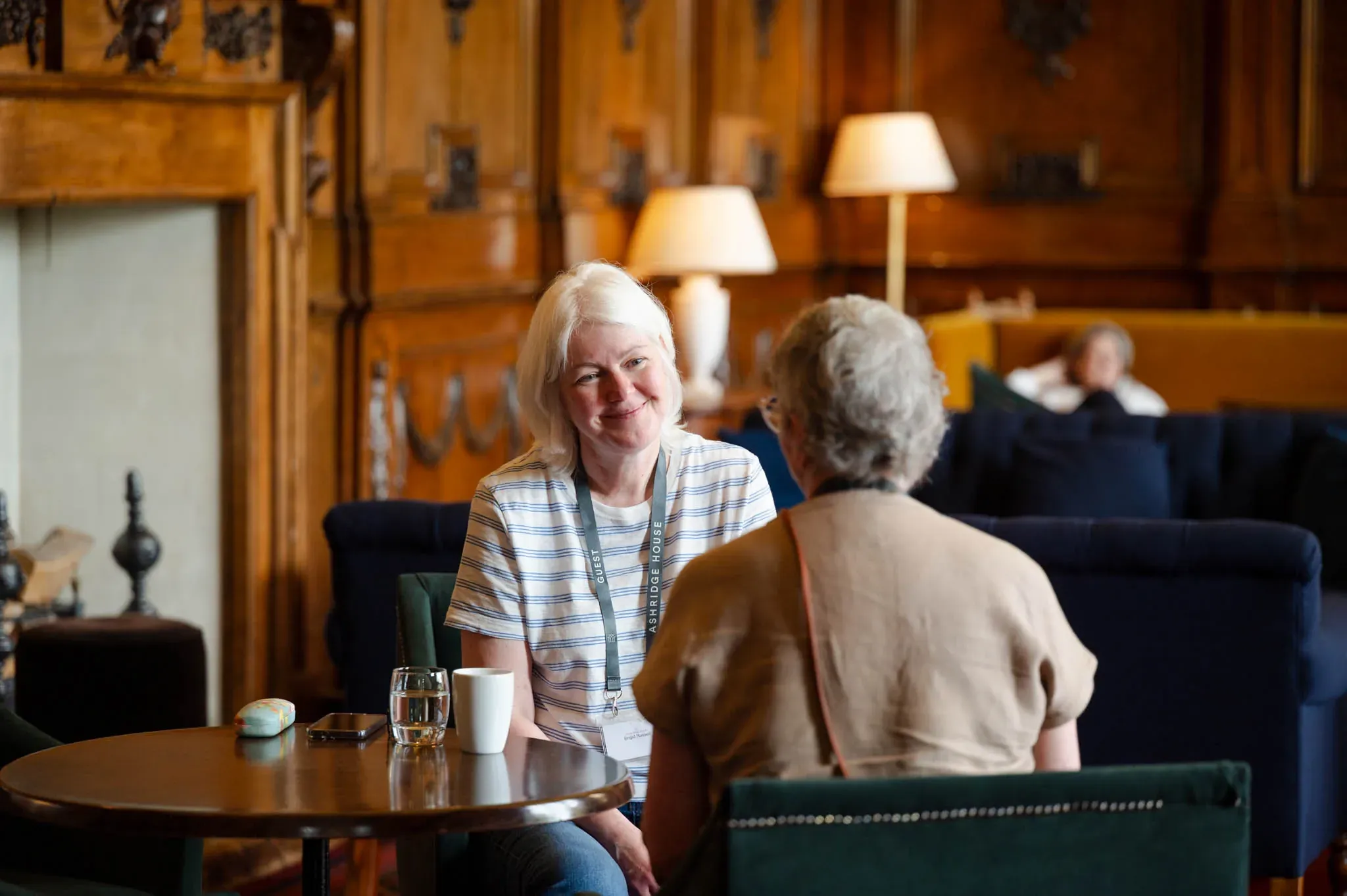 Two women sit at a wooden table in a cozy, wood-paneled room. One smiles warmly at the other, surrounded by comfortable seating and soft lighting.