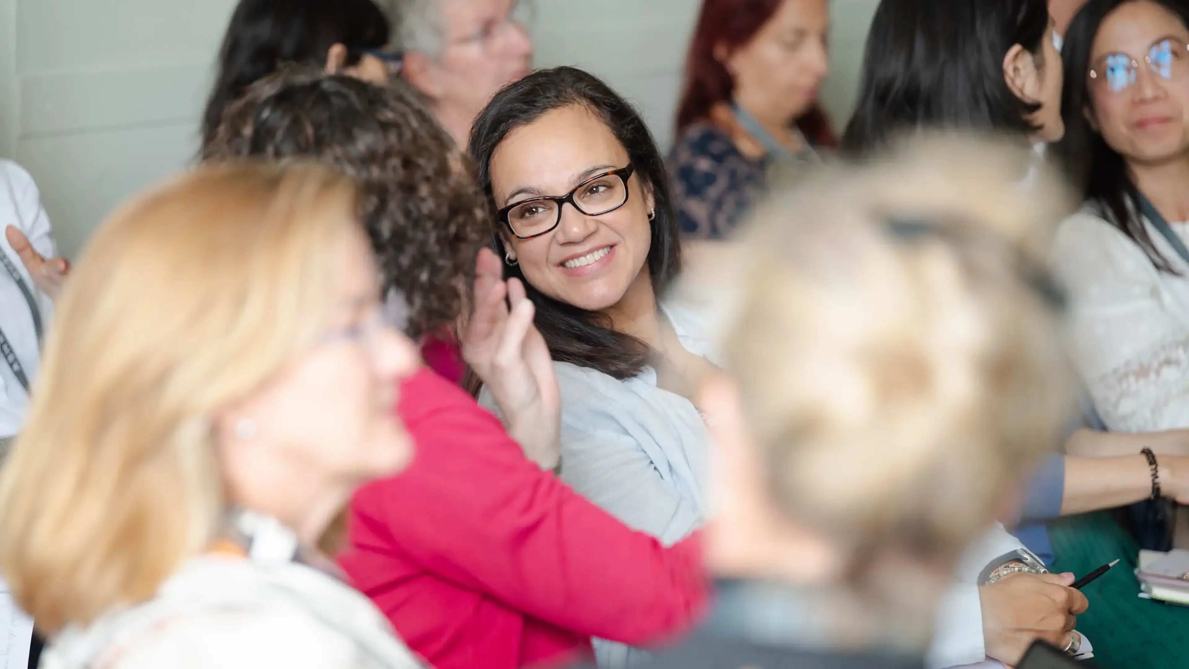 A smiling woman with glasses sits among a group of people, engaging in conversation.