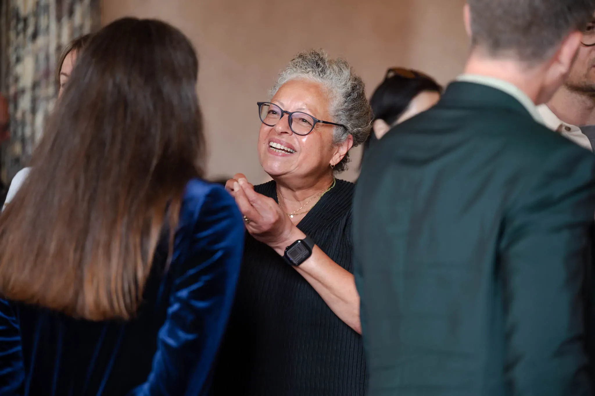 A smiling woman with glasses engages in conversation at a social event, surrounded by other people in semi-formal attire.
