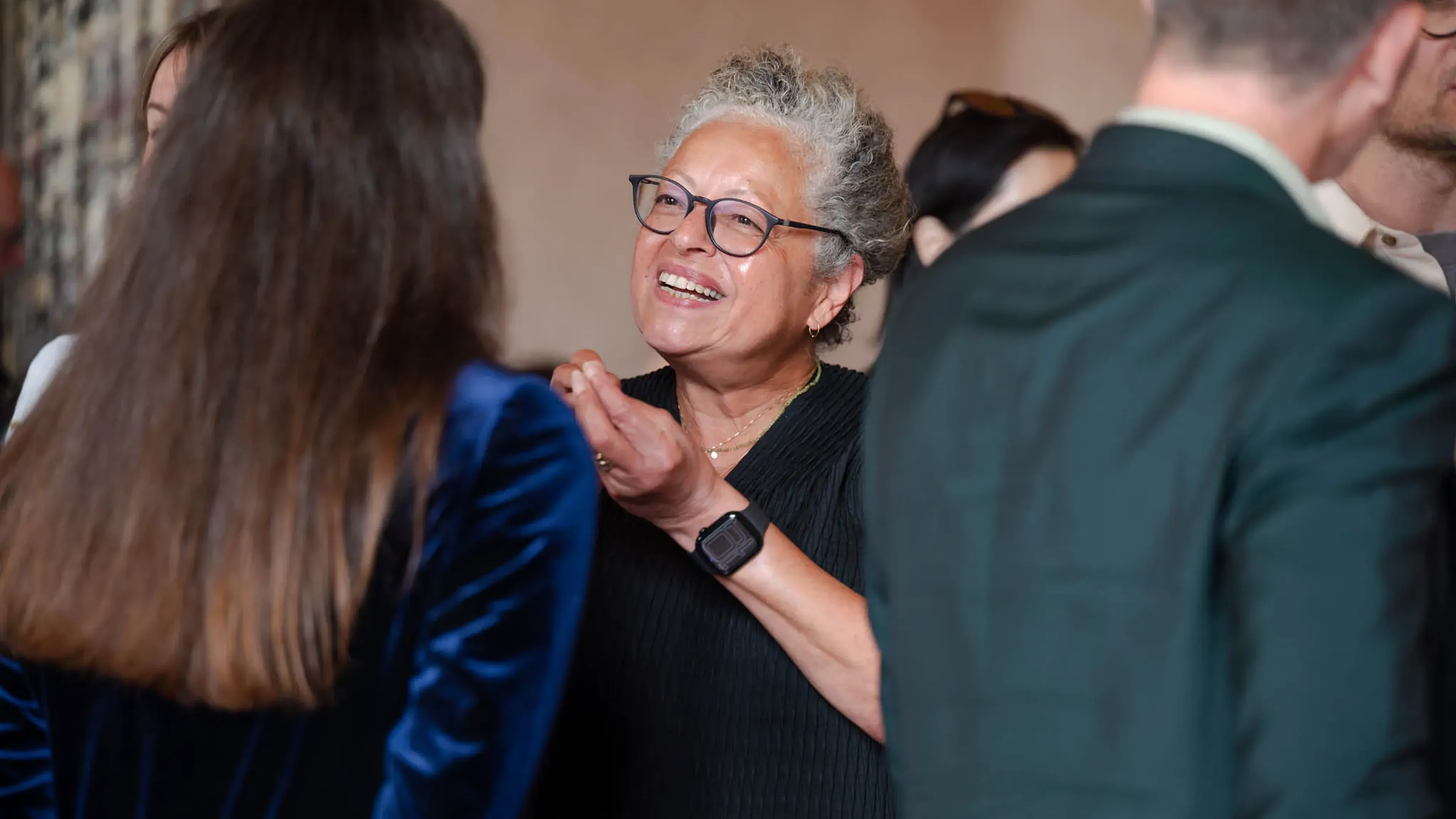 A smiling woman with glasses engages in conversation at a social event, surrounded by other people in semi-formal attire.