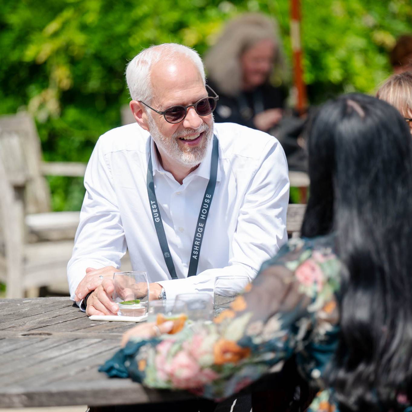 A man in sunglasses smiles while talking to two women outdoors at a wooden table.