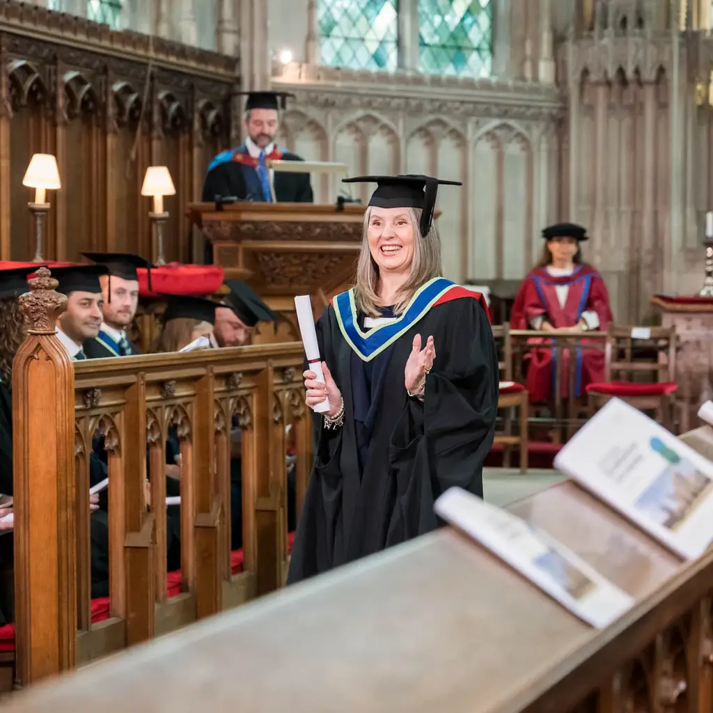 Graduate clapping with diploma in hand at a university ceremony in a historic hall.