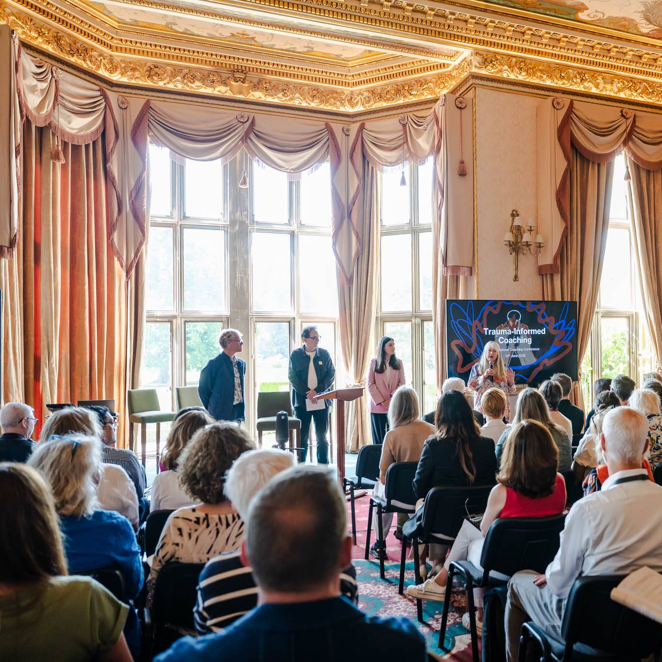 Audience attending a presentation on "Trauma-Informed Coaching" in an ornate room.
