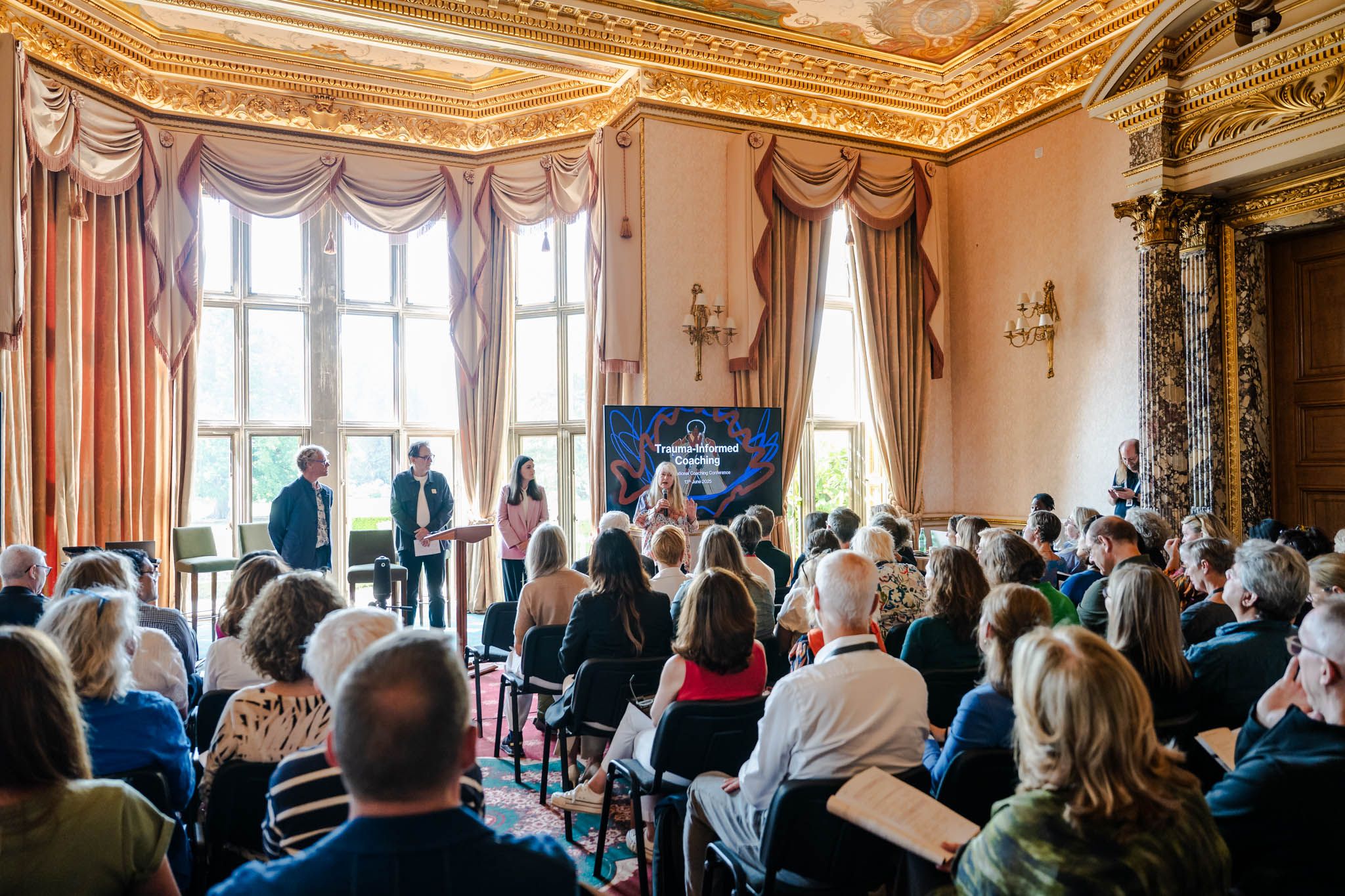Audience attending a presentation on "Trauma-Informed Coaching" in an ornate room.
