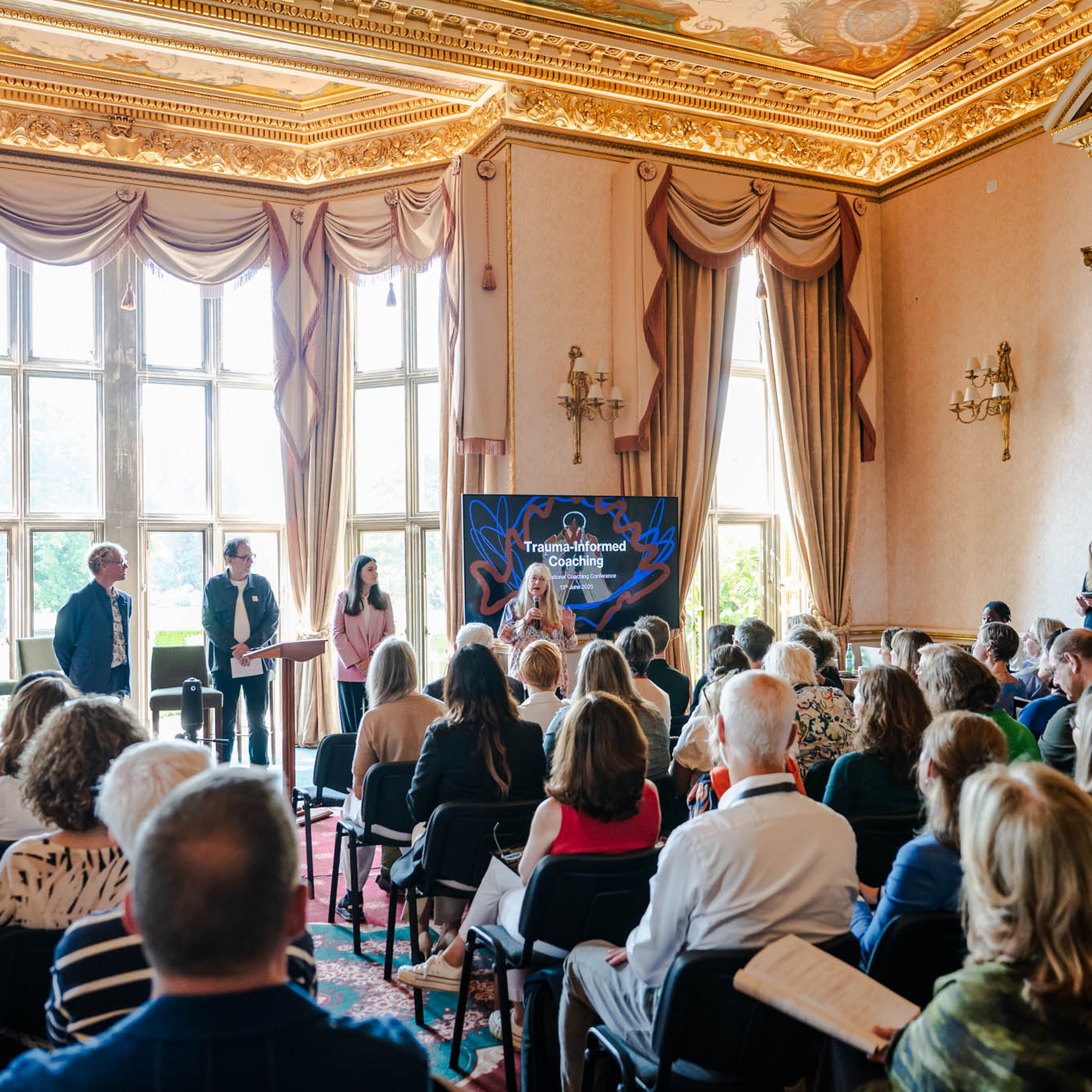 Audience attending a presentation on "Trauma-Informed Coaching" in an ornate room.