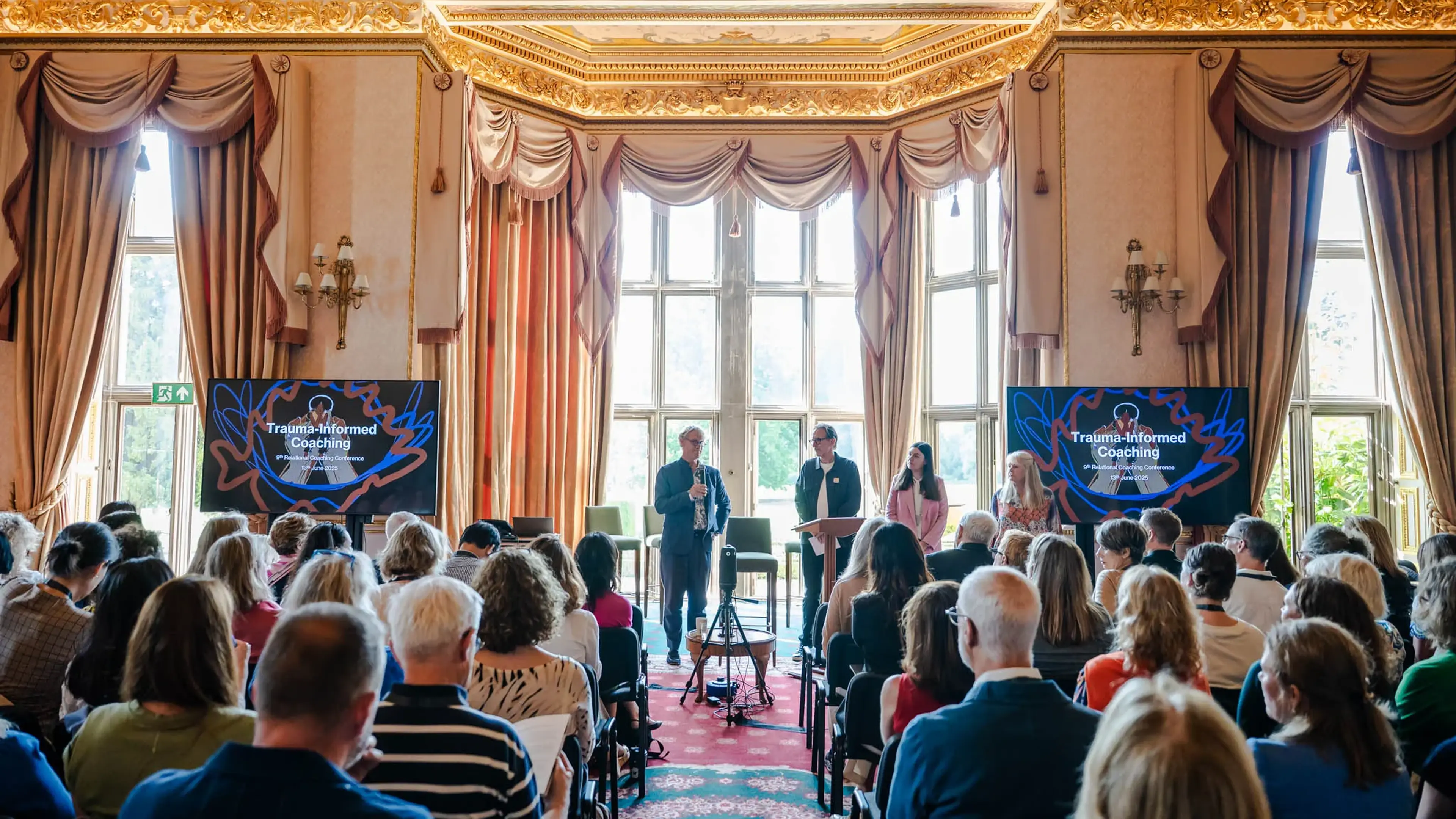 Audience listens to a "Trauma-Informed Coaching" presentation in a lavish room with large windows and ornate decor.