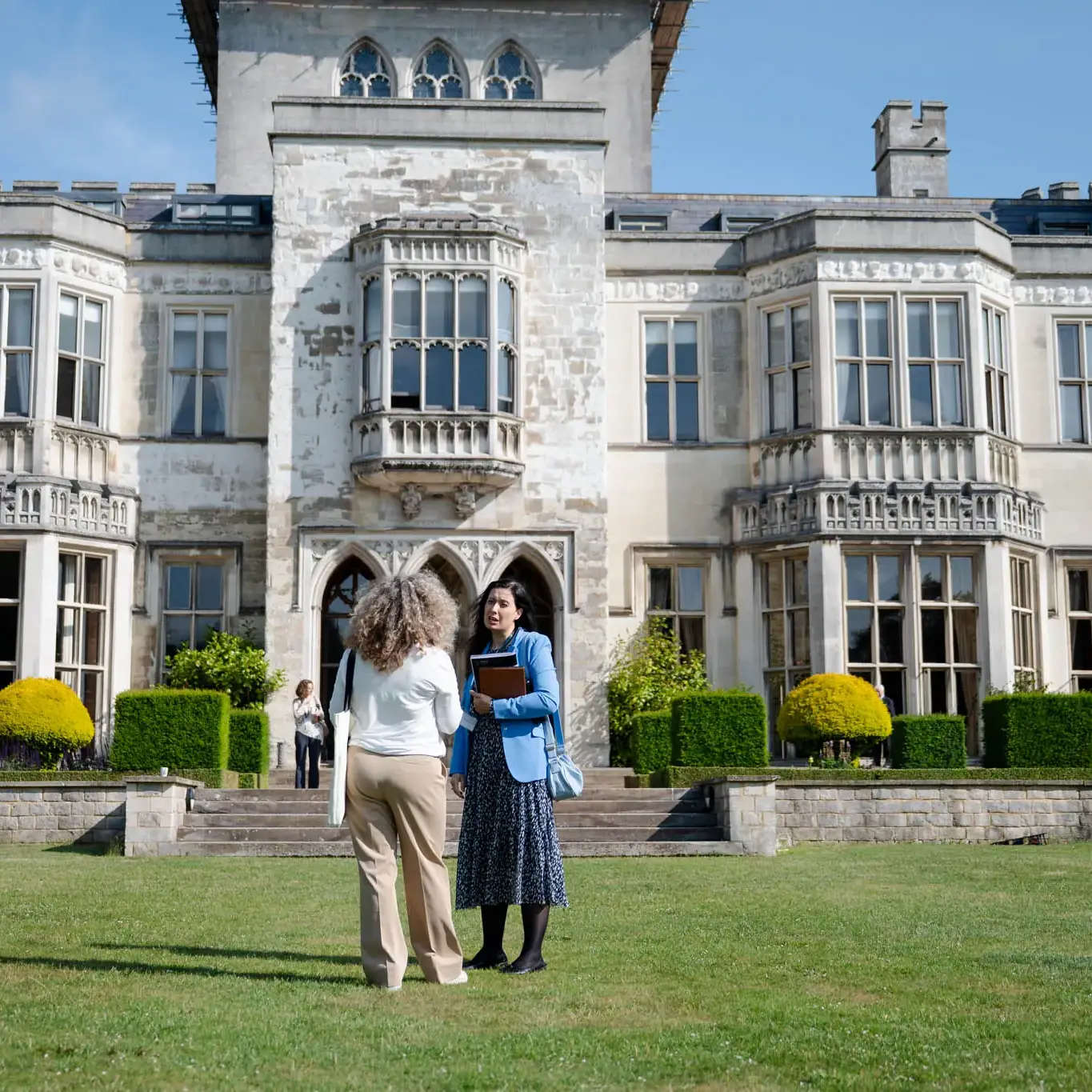 People converse in front of a historic stone mansion with arched windows and well-manicured gardens on a sunny day.