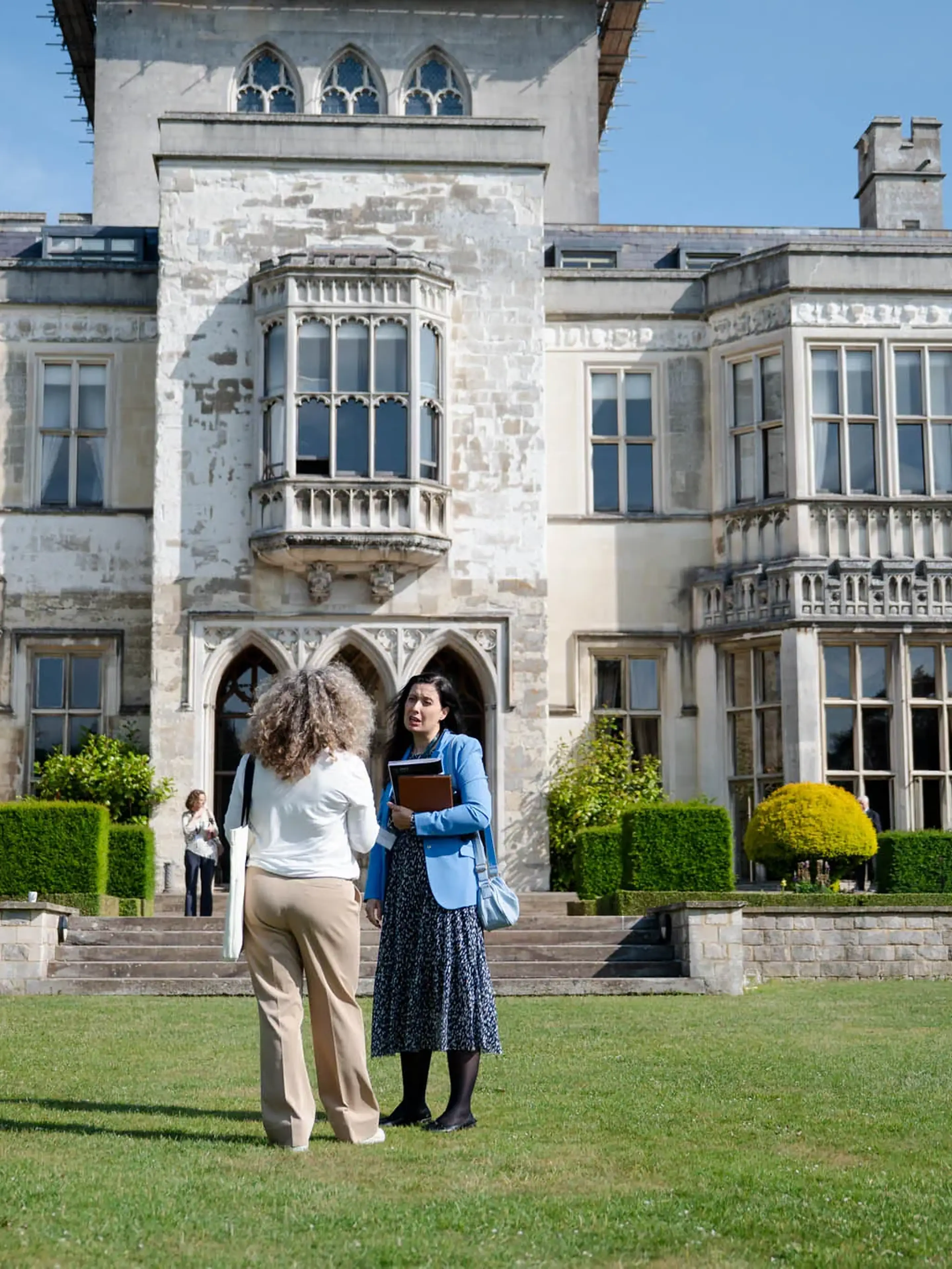 People converse in front of a historic stone mansion with arched windows and well-manicured gardens on a sunny day.
