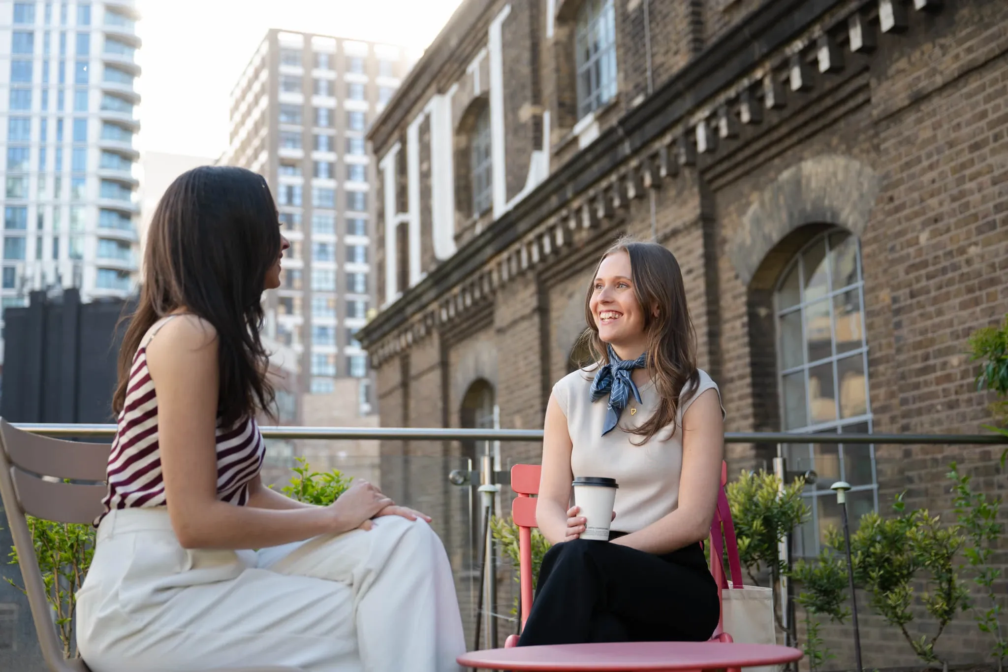 Two women sit and chat at an outdoor patio, one holding a coffee.