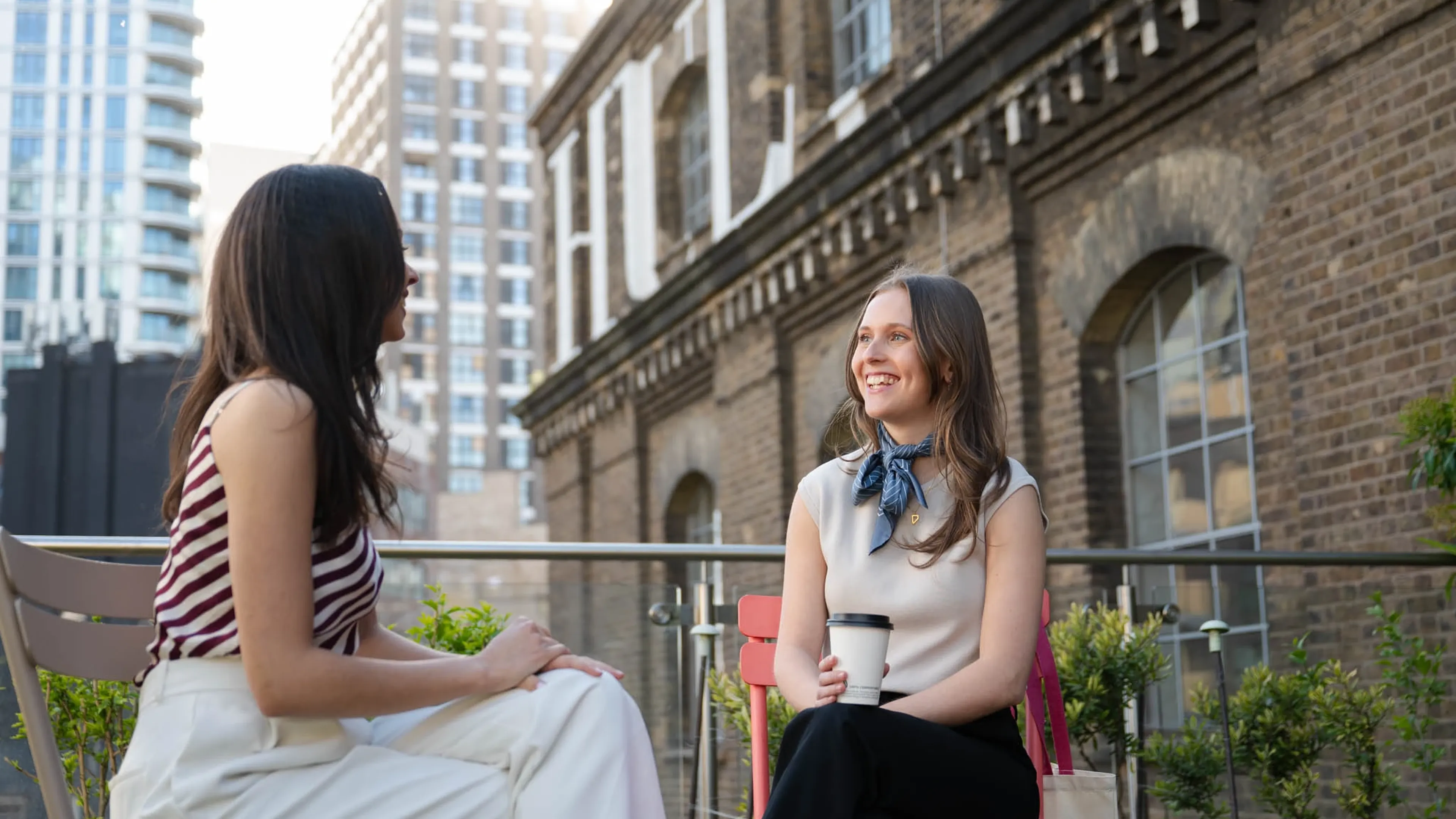Two women sit and chat at an outdoor patio, one holding a coffee.