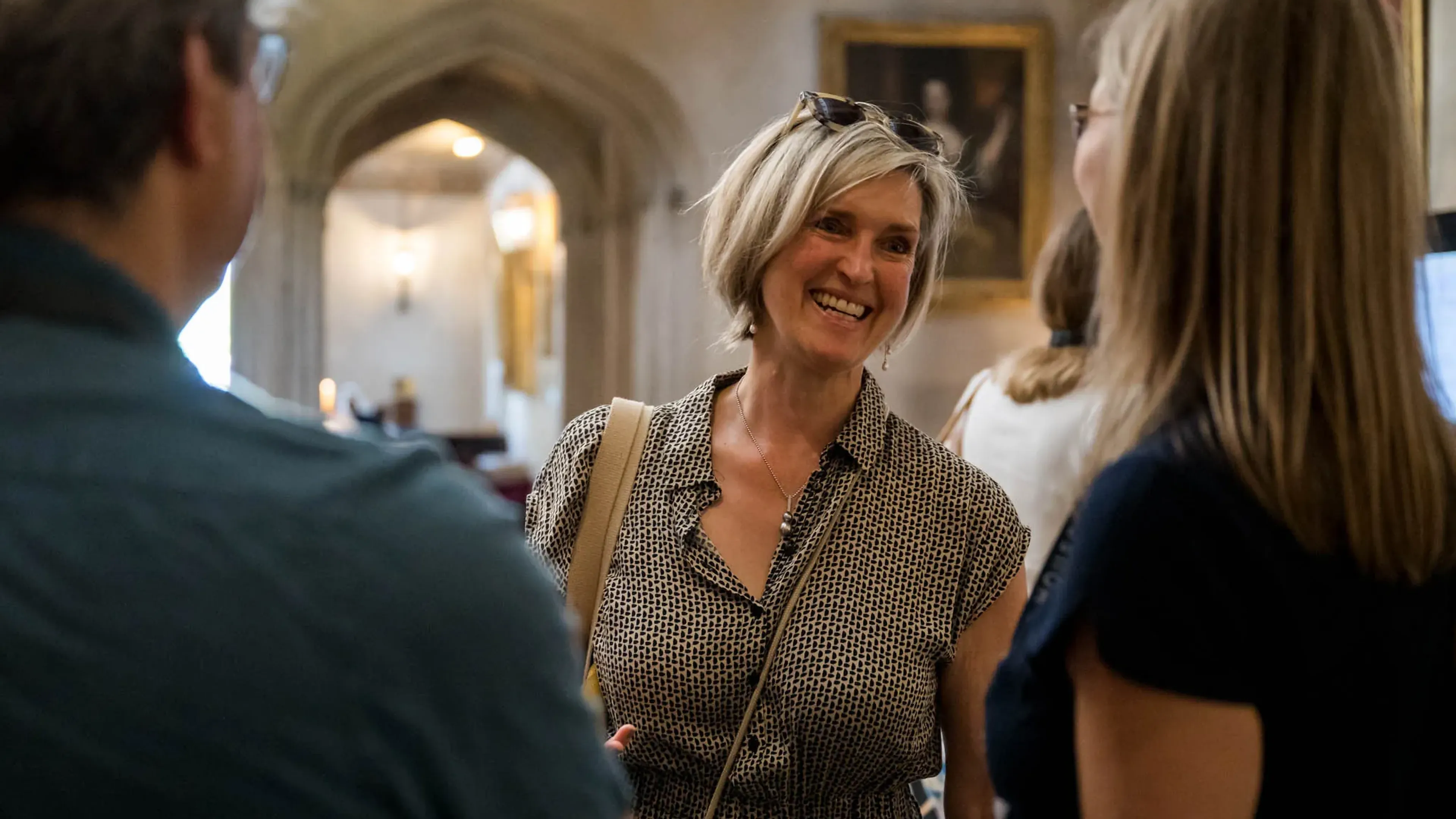 Smiling woman with sunglasses on her head converses with two people in a warmly lit room with arched doorways and framed portraits.