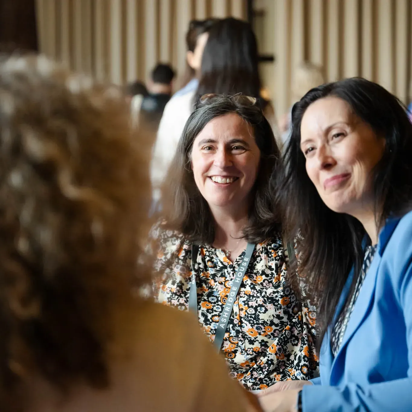 Two women sit smiling in conversation, surrounded by a group of people in a busy indoor setting.