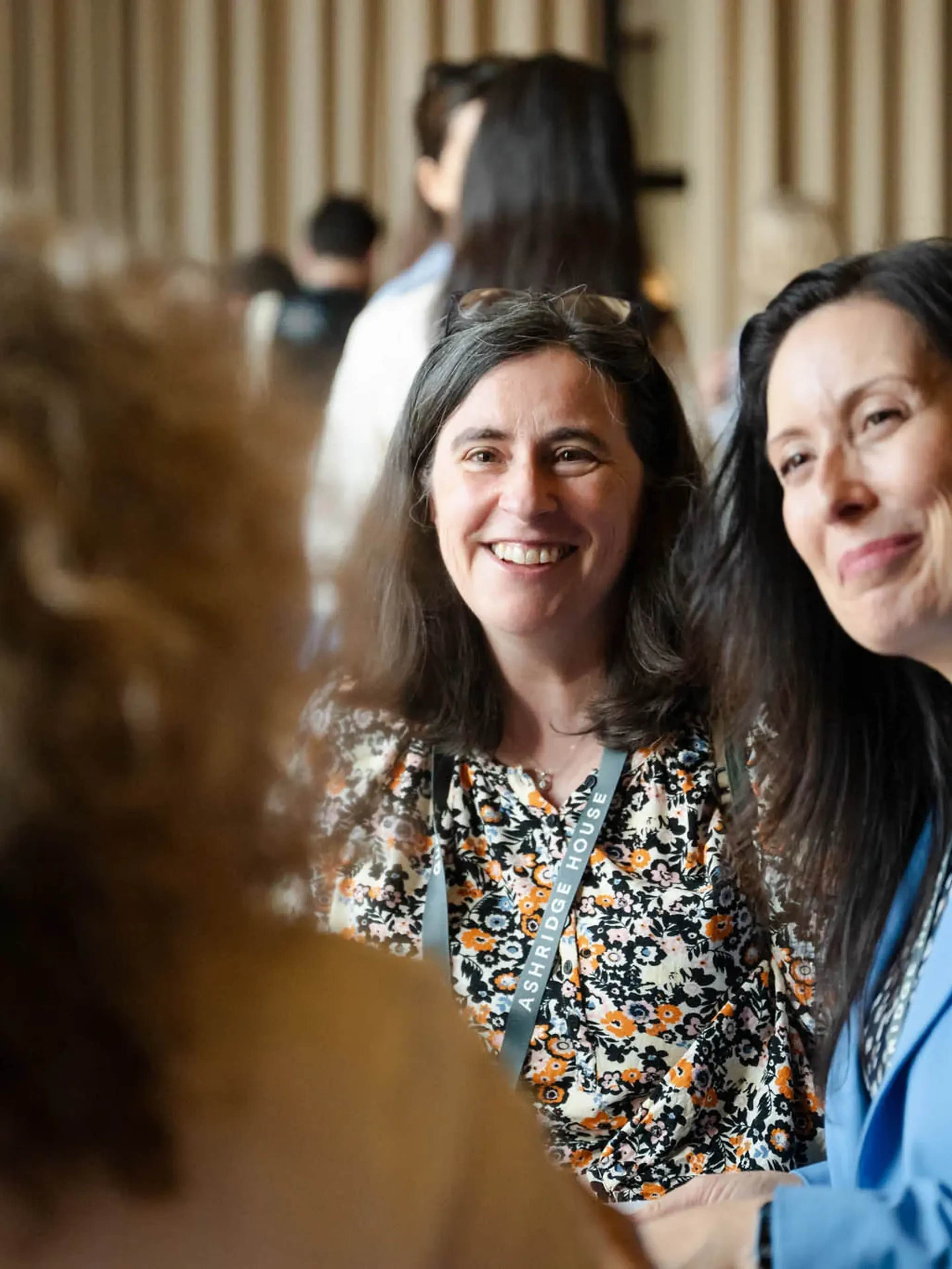 Two women sit smiling in conversation, surrounded by a group of people in a busy indoor setting.