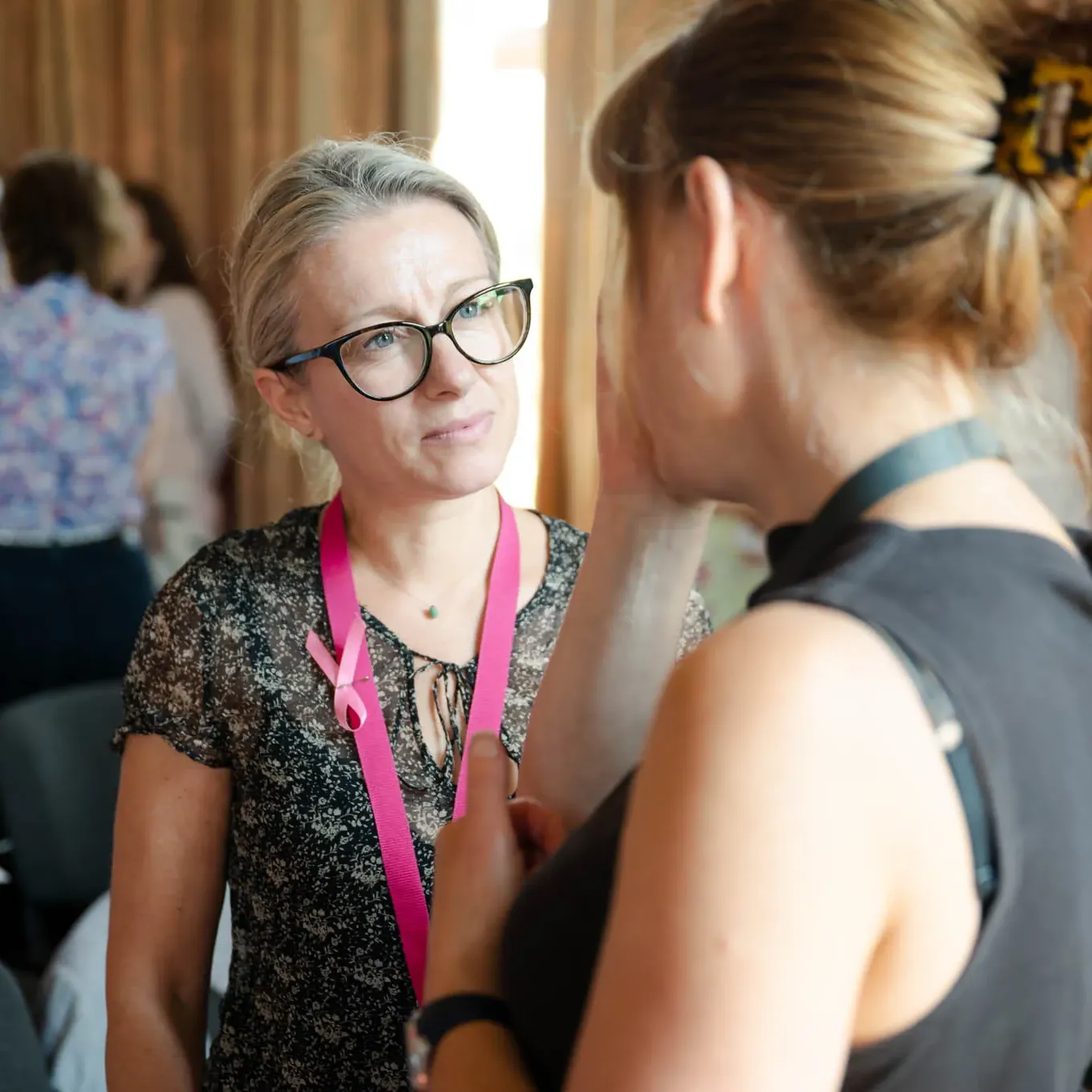 Two women engaging in conversation at an event, one wearing glasses and a pink ribbon lanyard.