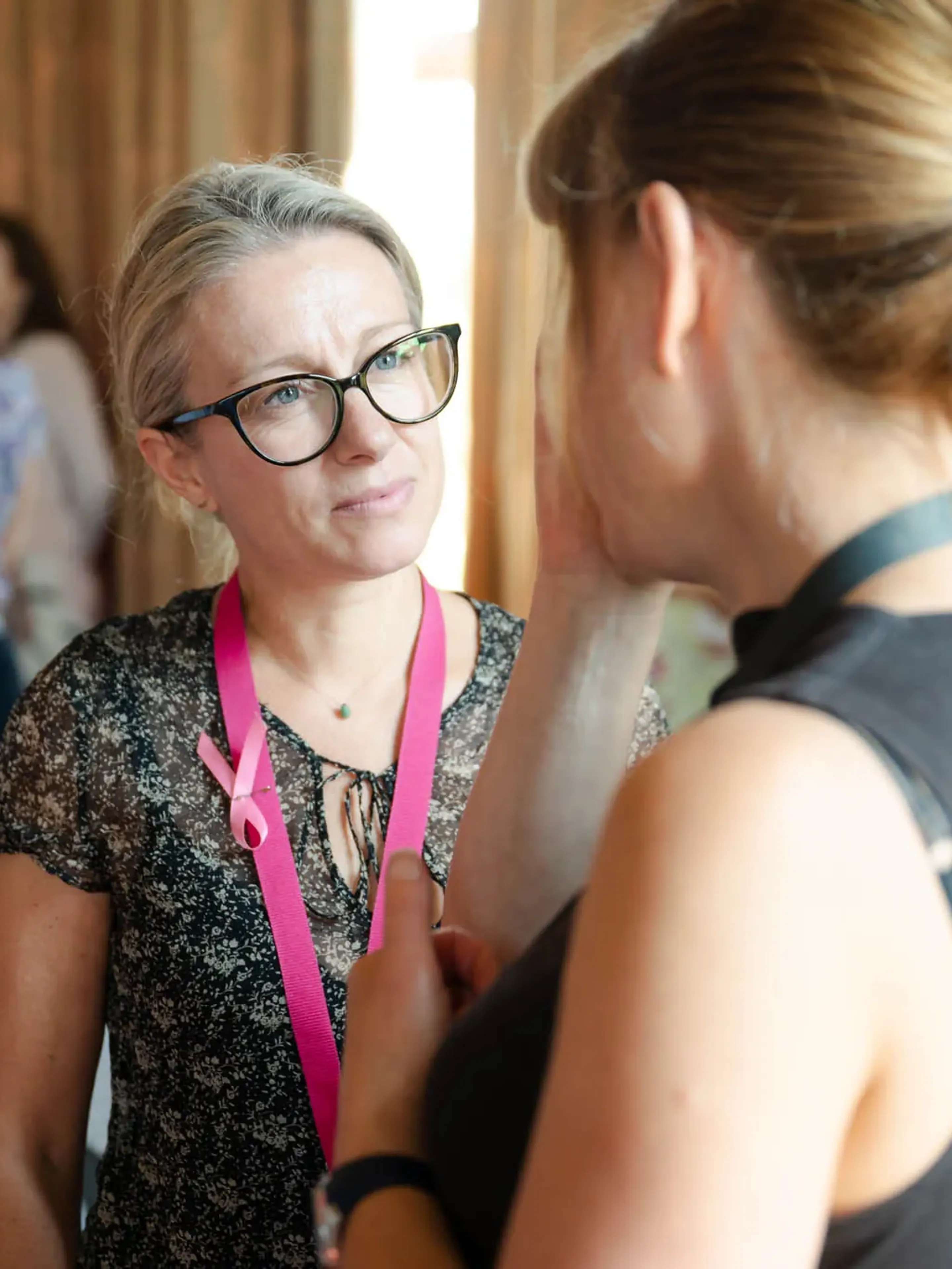 Two women engaging in conversation at an event, one wearing glasses and a pink ribbon lanyard.