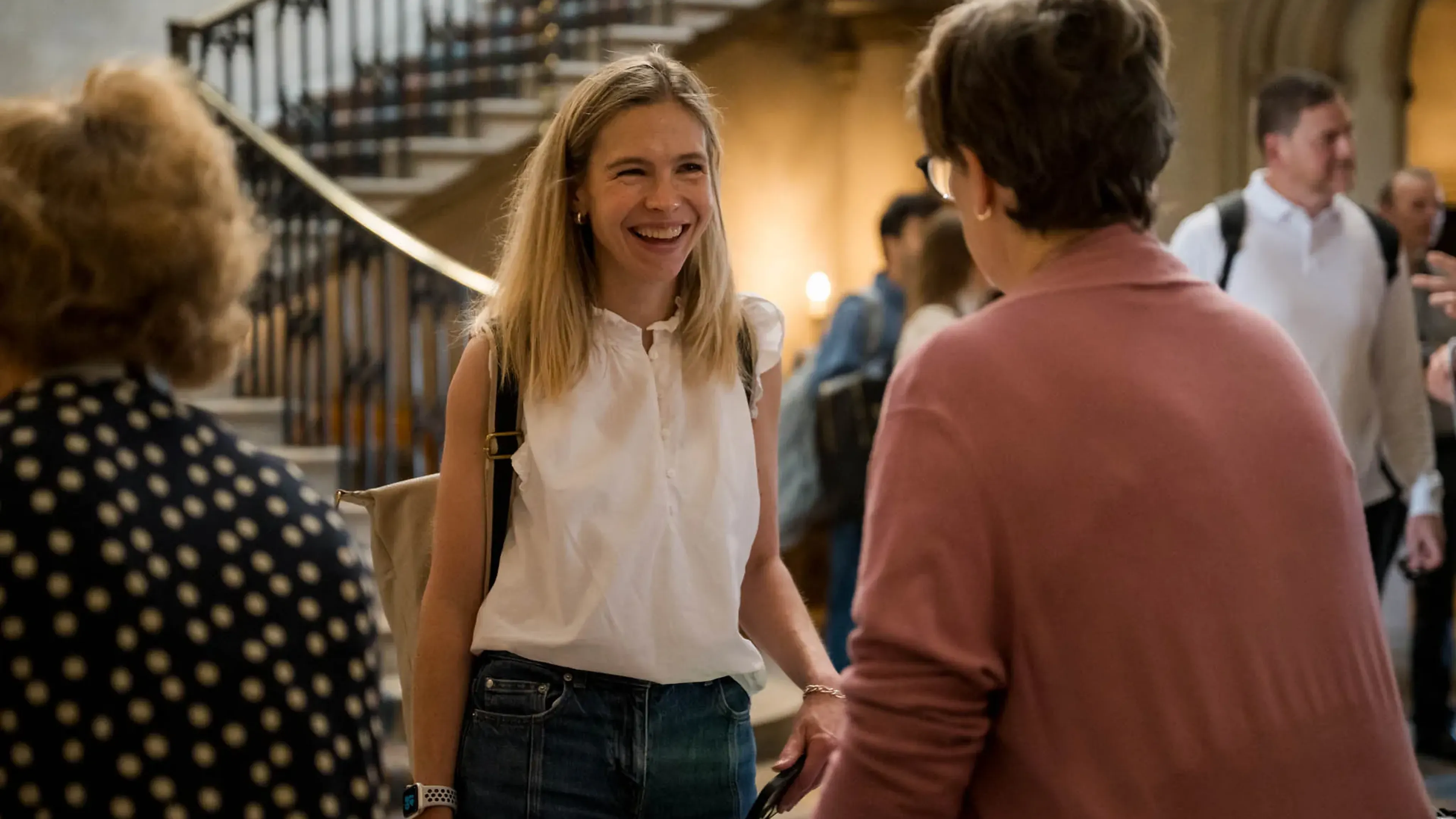 Two women conversing and smiling in a warmly lit room with a staircase in the background, surrounded by other people.