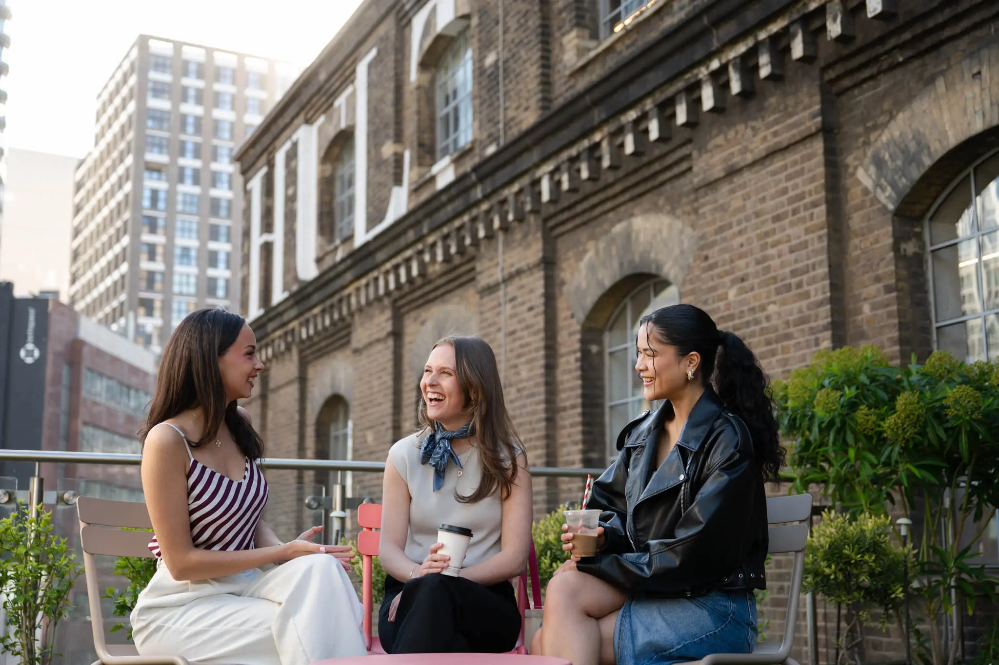 Three women laughing and chatting while holding drinks outside a brick building.