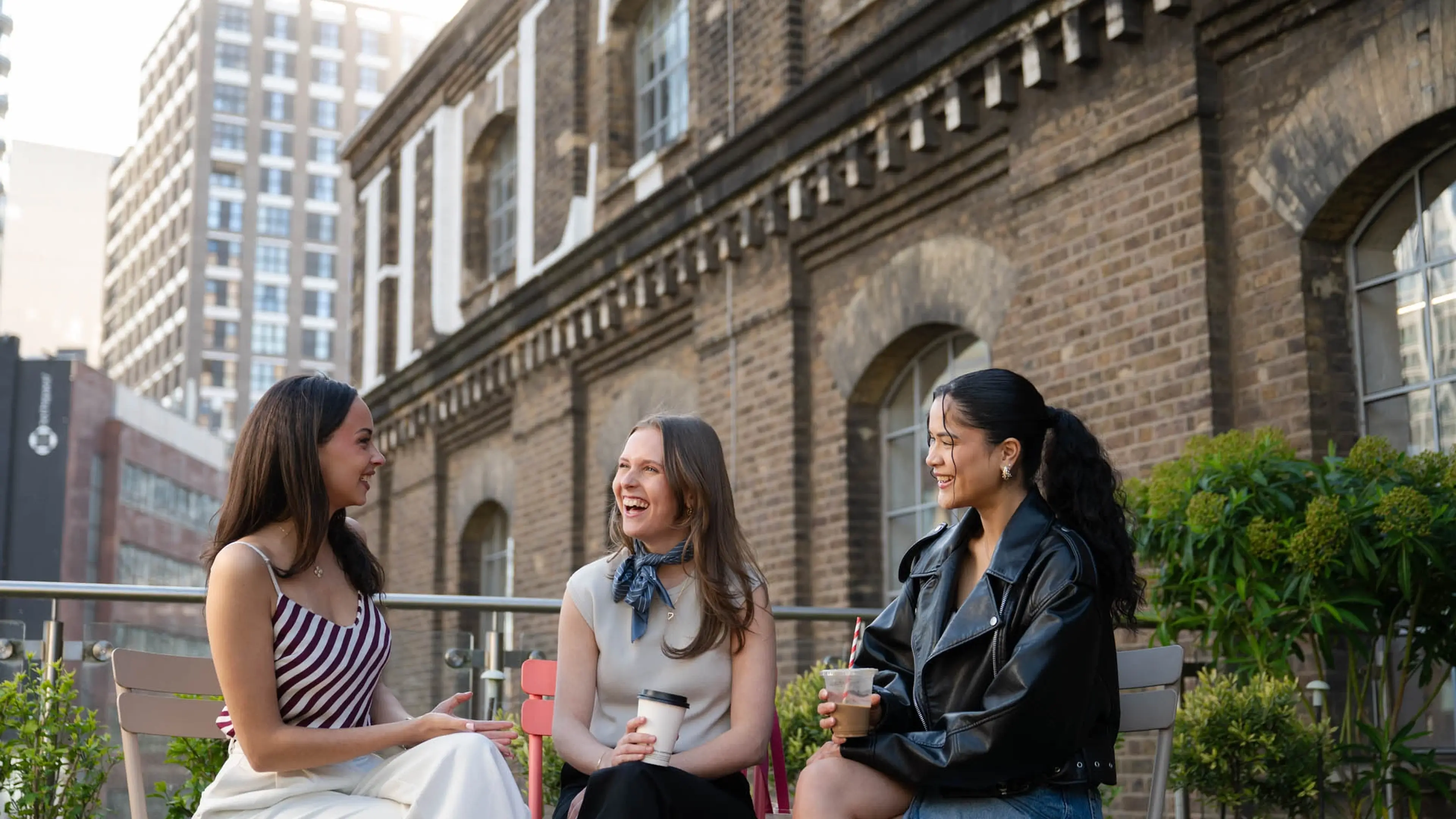 Three women laughing and chatting while holding drinks outside a brick building.