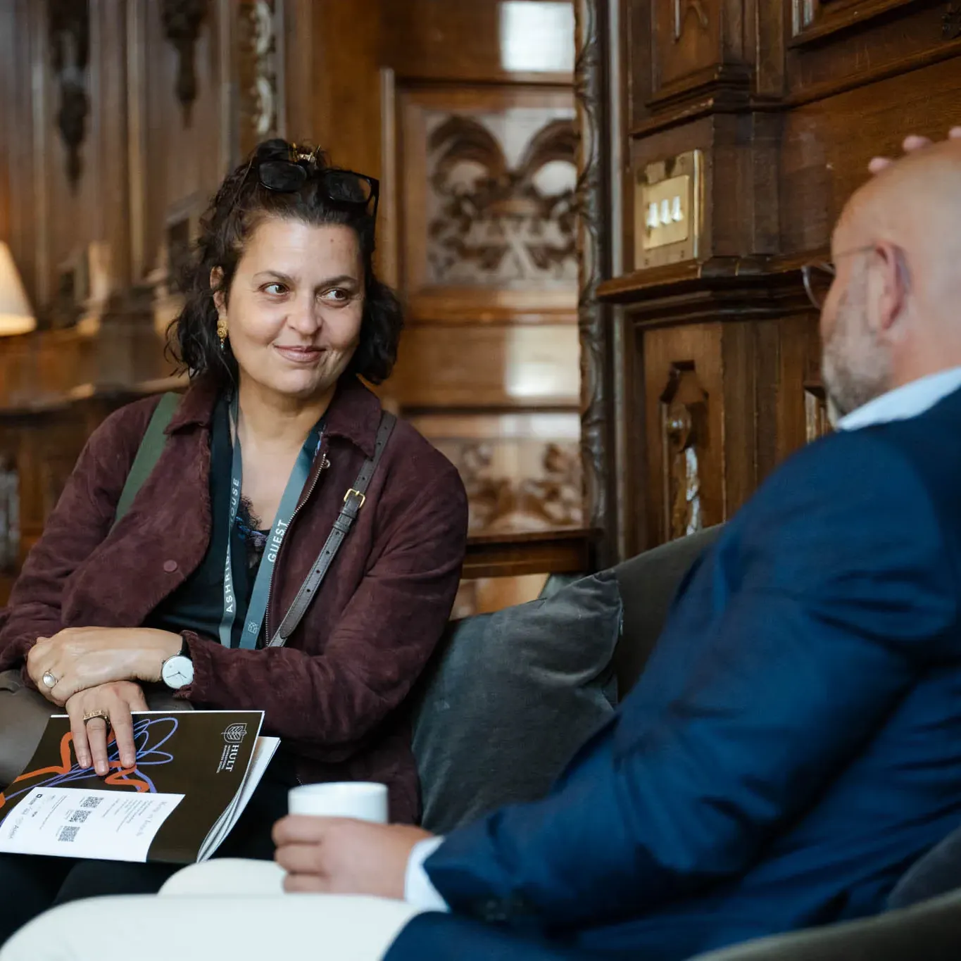 Two people engaged in conversation in a wooden-paneled room, one holding a booklet, the other a cup, with a lamp in the background.
