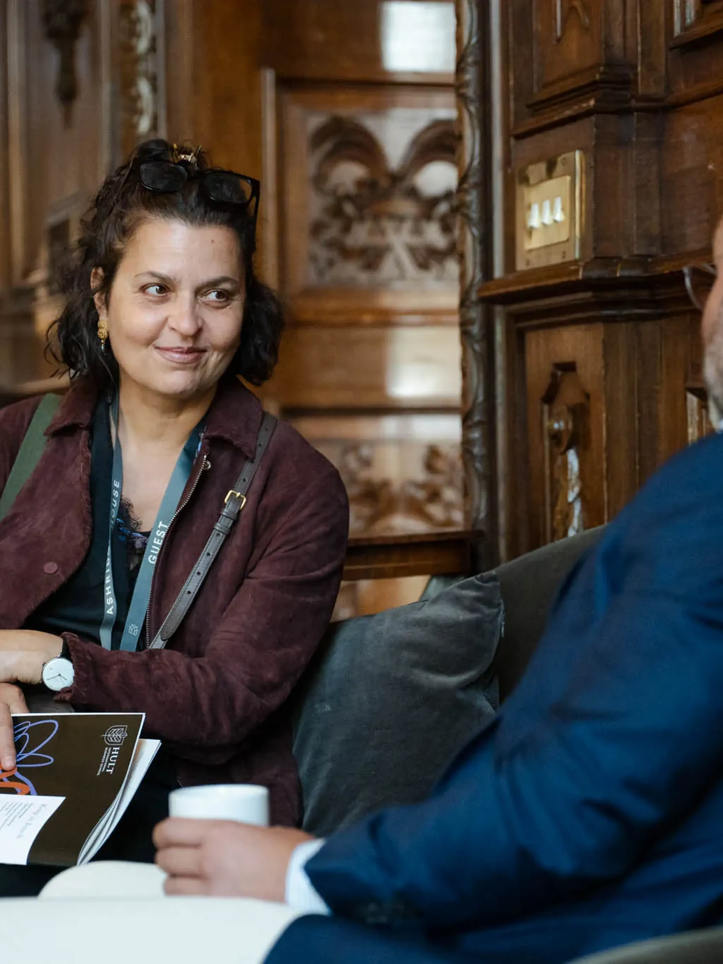Two people engaged in conversation in a wooden-paneled room, one holding a booklet, the other a cup, with a lamp in the background.