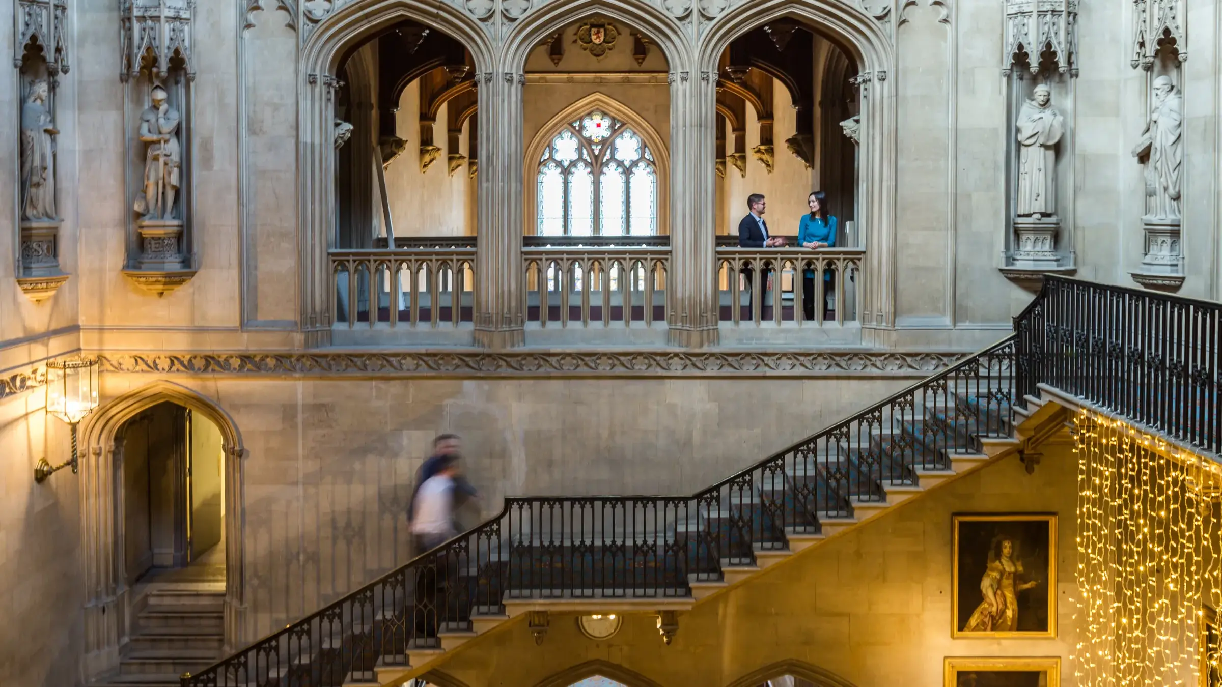 Gothic-style interior with ornate arches, two people conversing on the upper balcony, a blurred figure on stairs, and illuminated portraits below.