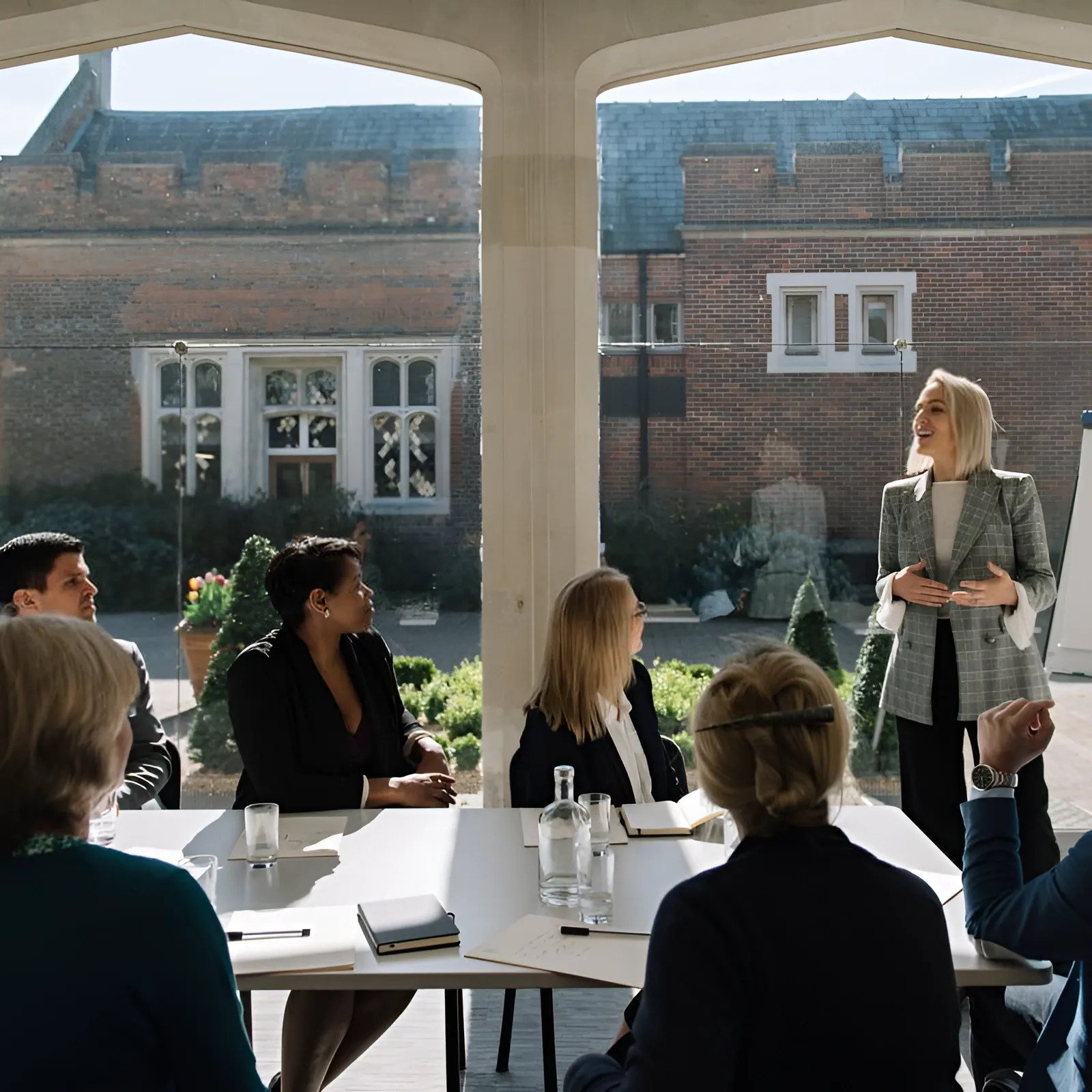 A woman presents to a group of people seated at a table in a bright room.