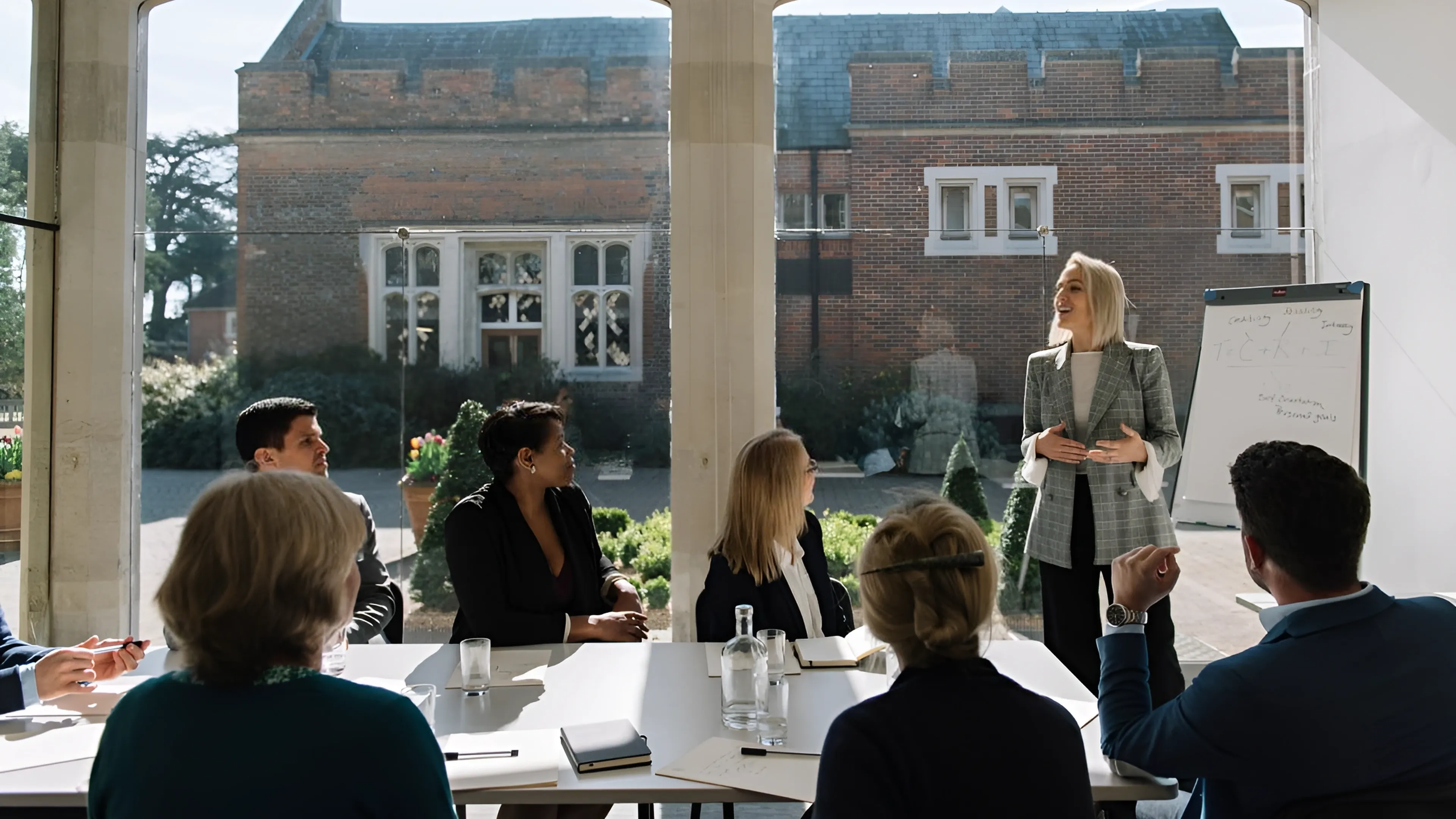 A woman presents to a group of people seated at a table in a bright room.