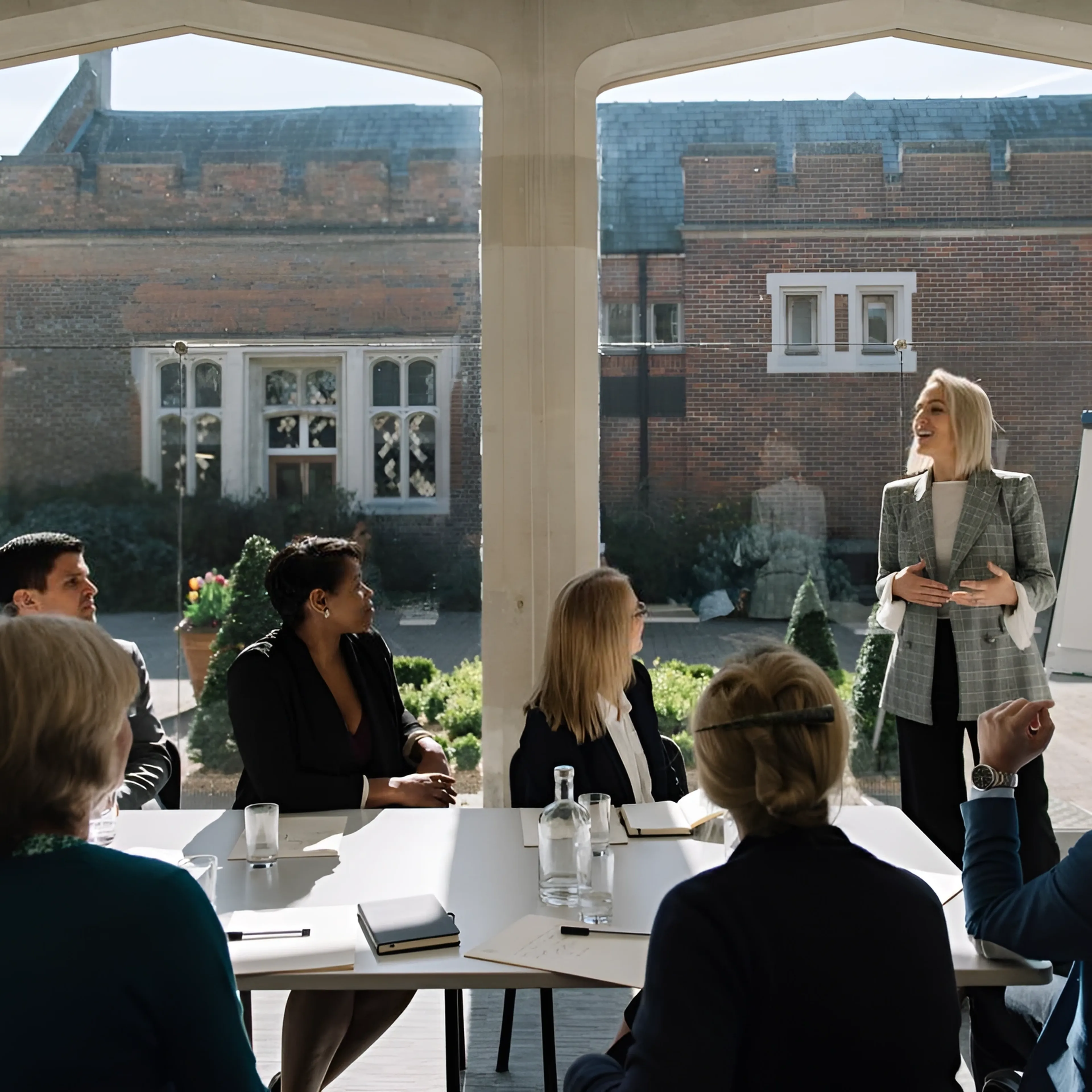A woman presents to a group of people seated at a table in a bright room.