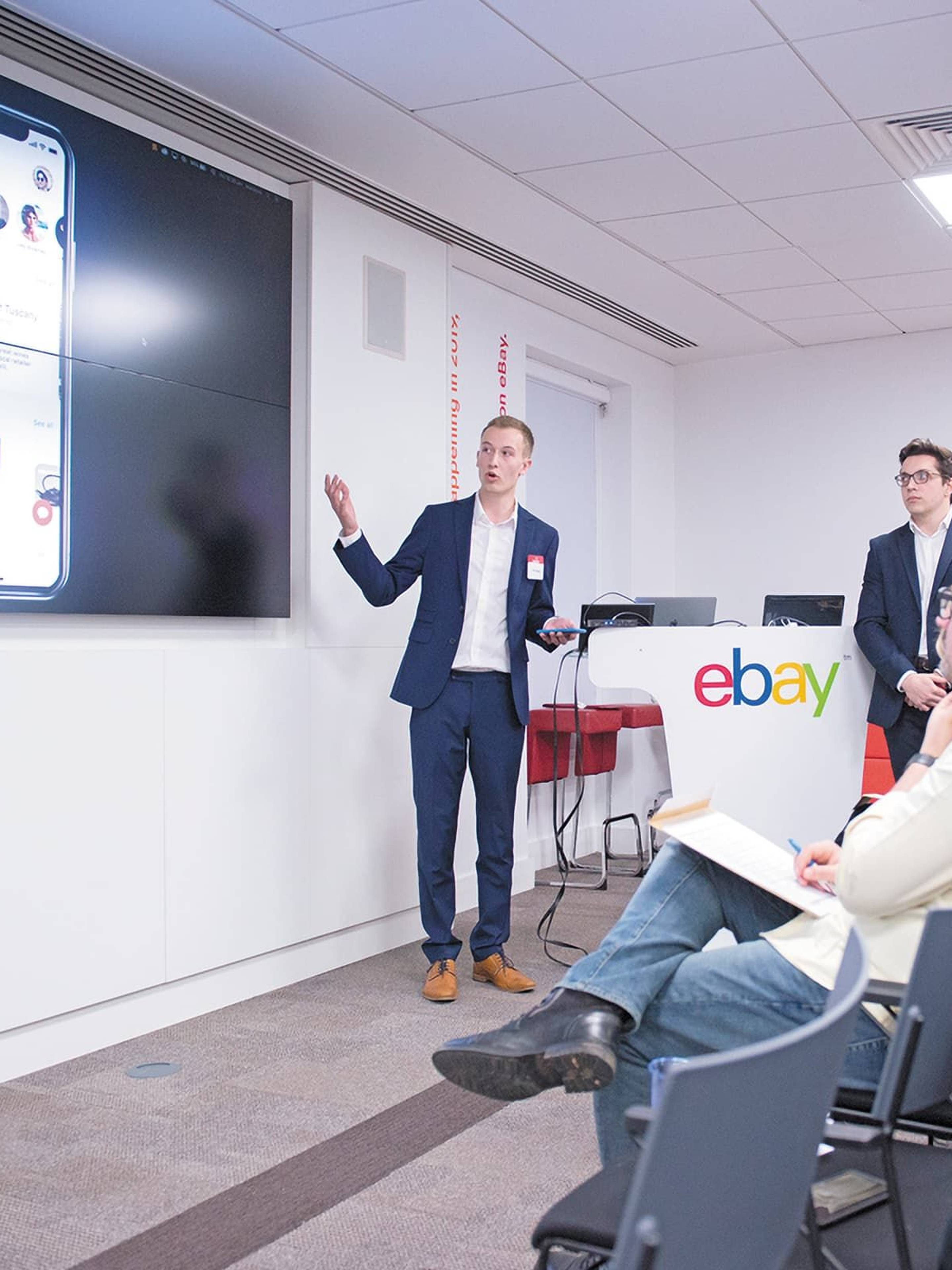 Two men present to an audience in a conference room with eBay logos.