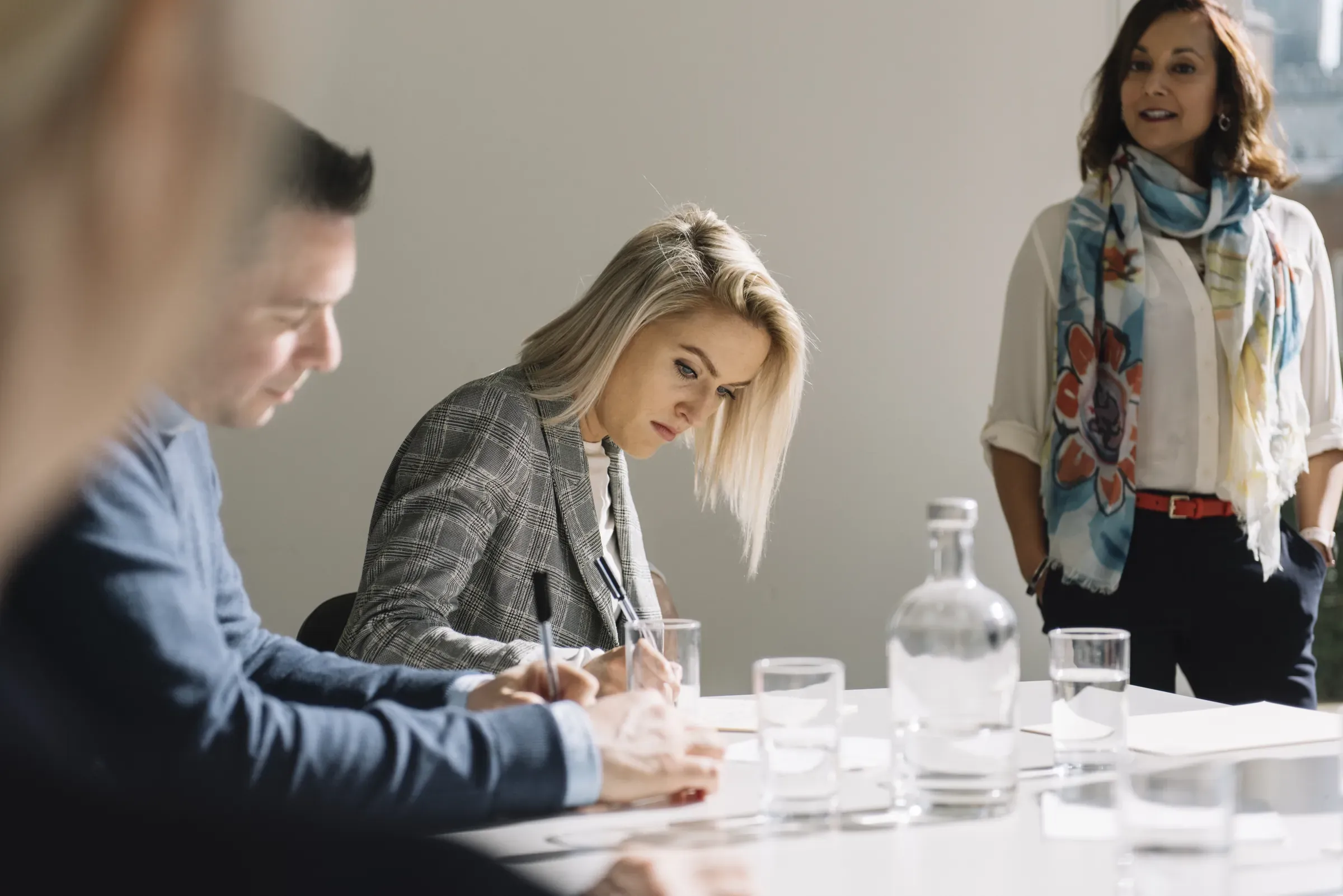 People in a meeting room, one woman writing notes, another standing, and a man listening, with water glasses on the table.