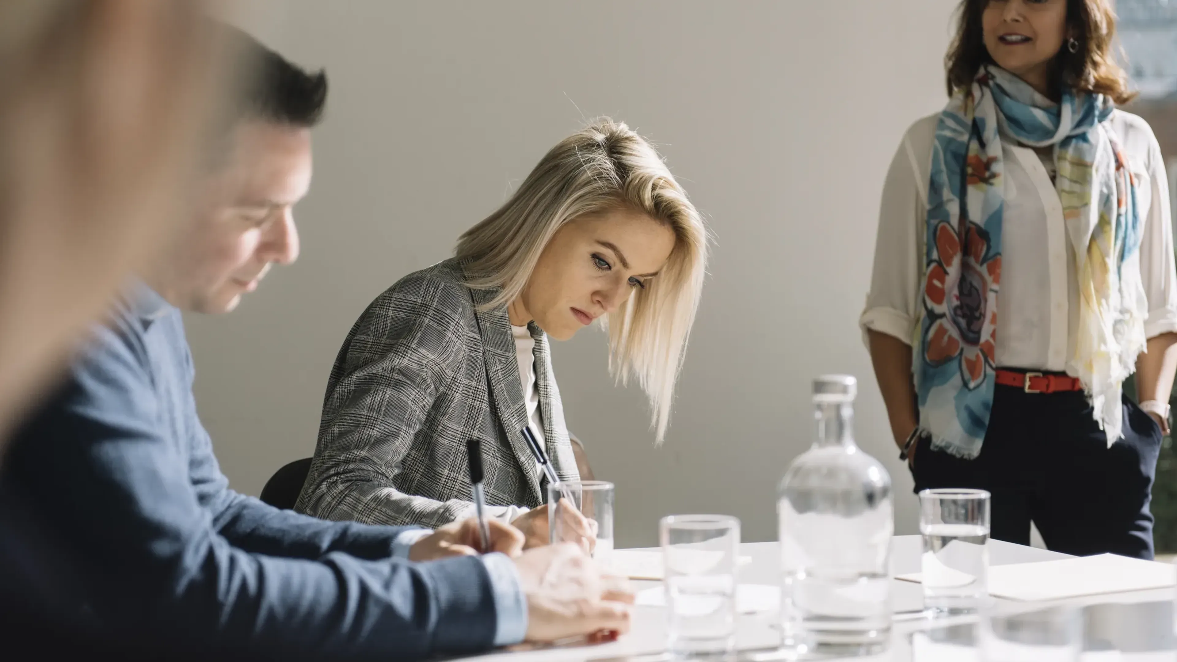 People in a meeting room, one woman writing notes, another standing, and a man listening, with water glasses on the table.