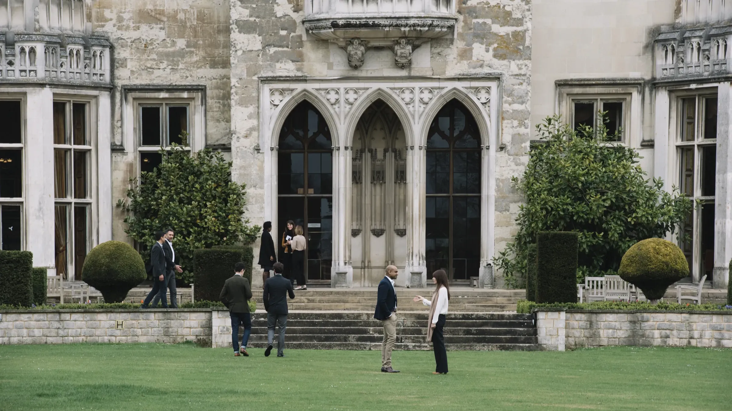 People socialize outside a historic stone building with arched doors, surrounded by manicured greenery and a lush lawn.