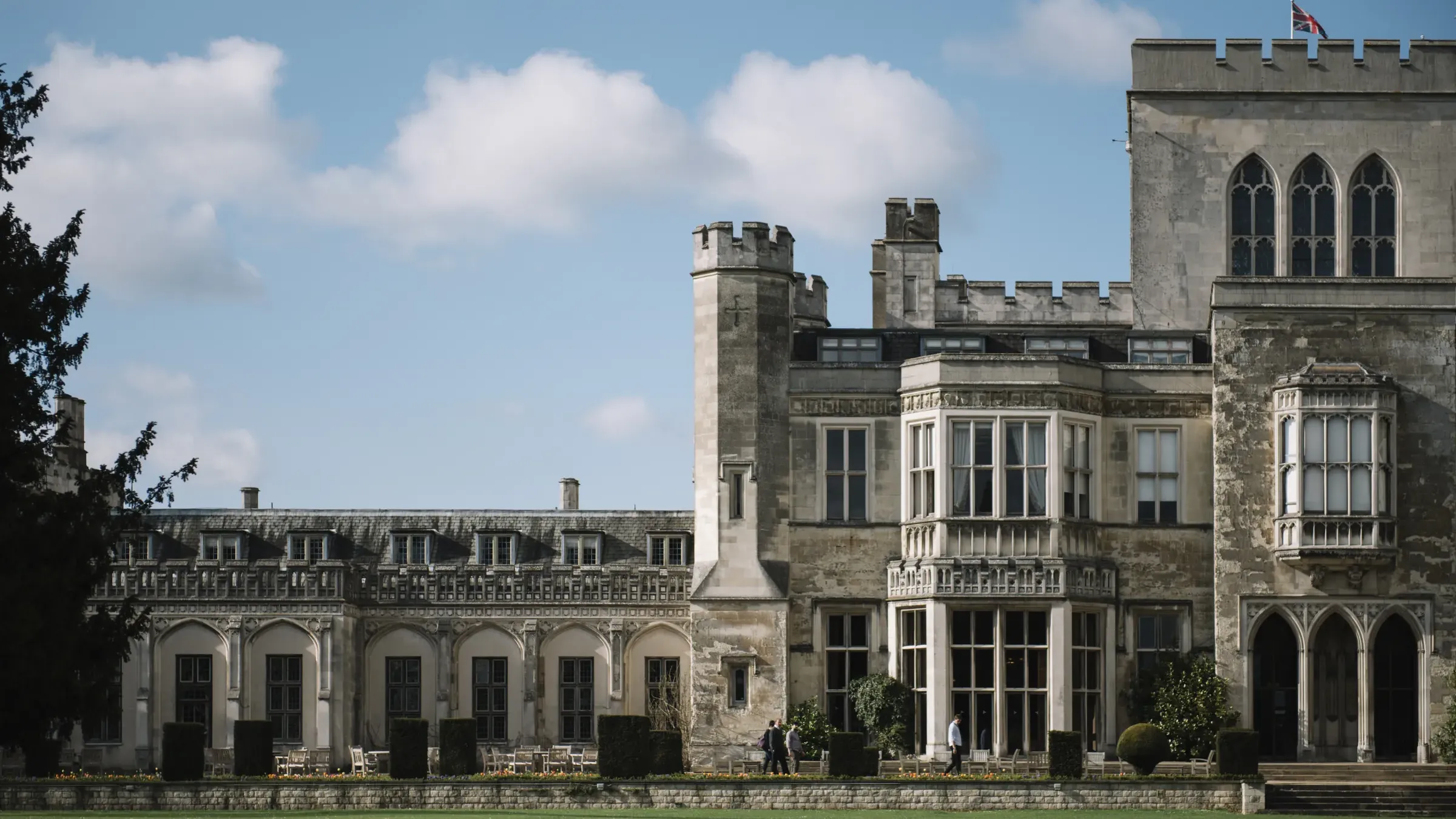 Historic stone building with ornate windows and turrets under a blue sky with clouds. People walking in the garden.